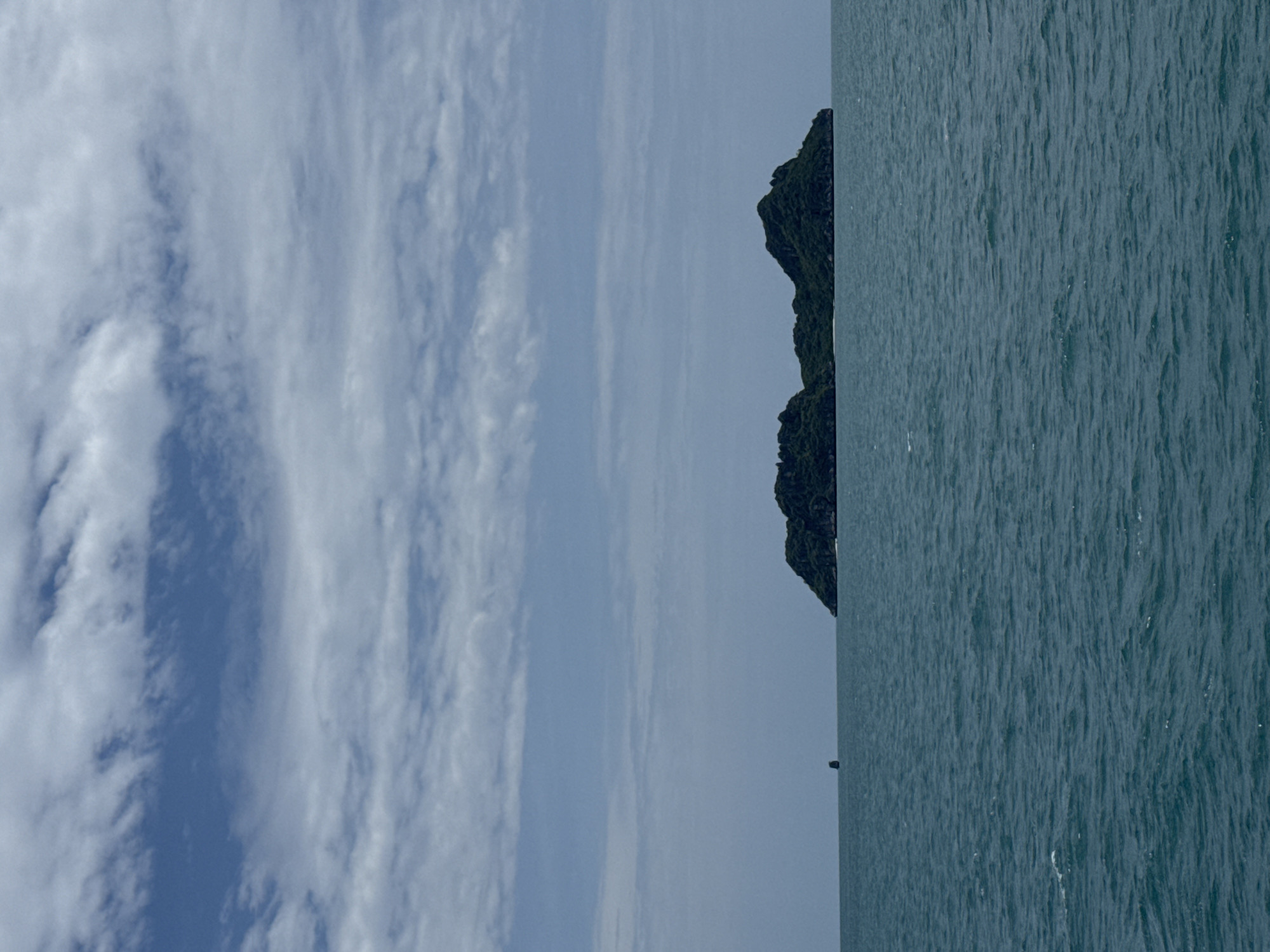 The image depicts a calm, blue ocean with a small, tree-covered island in the distance. The sky is partly cloudy with white, wispy clouds against a pale blue background. The island appears rugged and uninhabited, with a lush green canopy. There is a very small dark object, possibly a boat or buoy, visible in the water to the left of the island. The water's surface is slightly rippled. The scene conveys a sense of tranquility and isolation. This appears to be a view of Koh Tao, an island located northwest of Ko Pha-ngan, Thailand, known for its clear waters and diving sites.