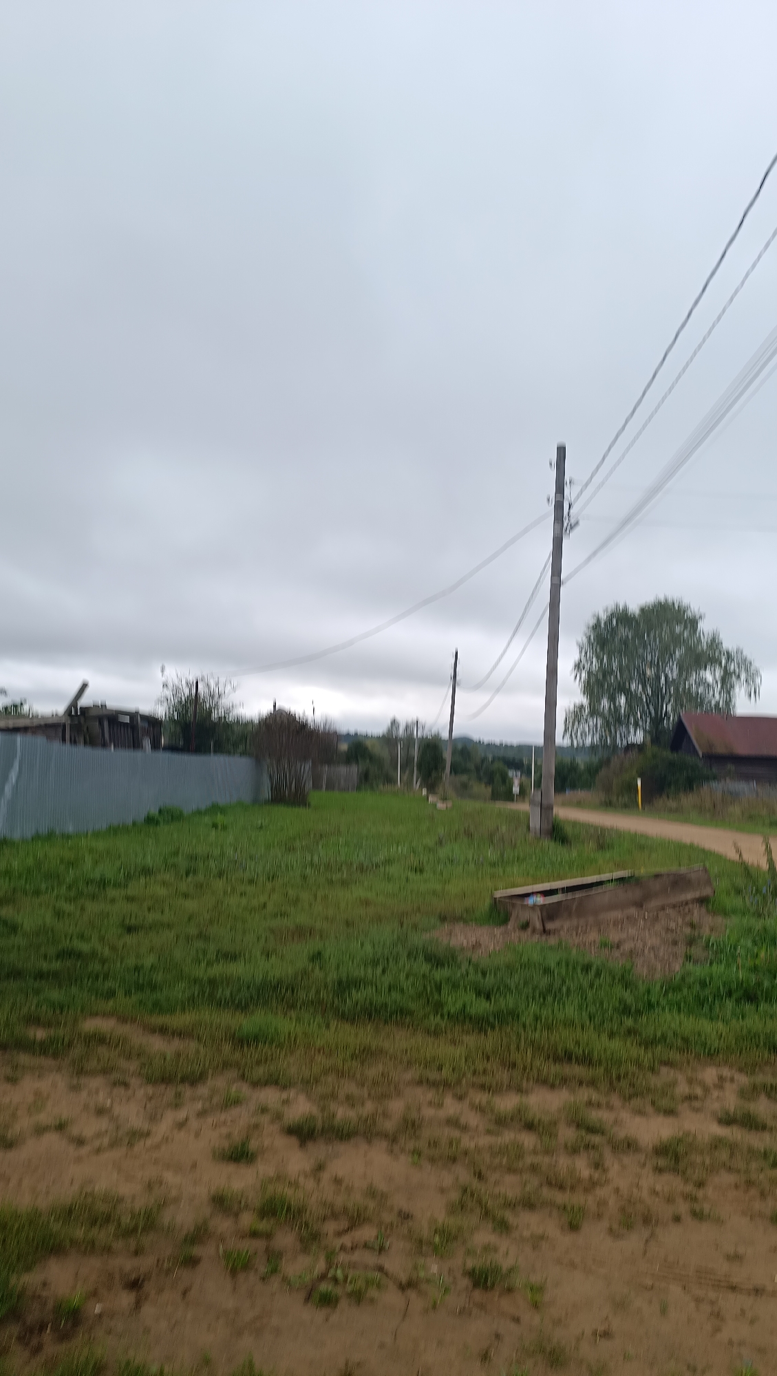 The image shows an overcast day in a rural area, possibly Karsha, Russia. The scene is dominated by a grassy field with a dirt path winding through it. In the foreground, there is a concrete trough filled with some debris. Further back, a wooden fence borders the field, with some rustic structures visible beyond it. Power lines stretch across the sky, supported by wooden poles, a common sight in rural Russian landscapes. A large tree with green foliage stands to the right, near a building with a reddish roof. The overall impression is one of a quiet, undeveloped village on a cloudy day.