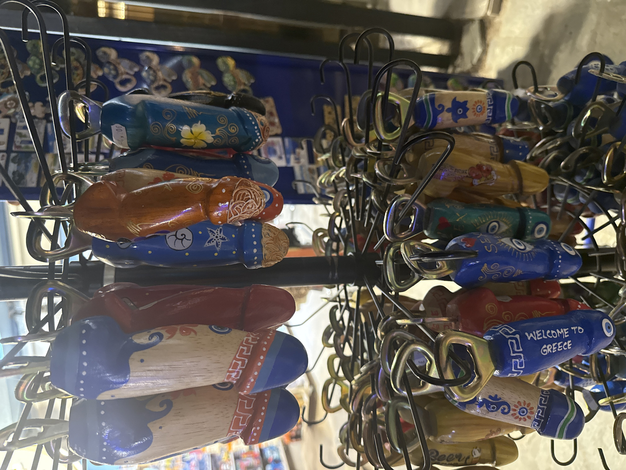 The image displays a close-up of a display rack filled with numerous phallic-shaped bottle opener keychains at a souvenir shop in Ofrynio Beach, Greece. The keychains are made of wood and painted with various designs, including Greek patterns, flowers, evil eyes, and the text "WELCOME TO GREECE." The keychains are arranged in tiers, with the top tier featuring larger, more detailed items and the lower tiers filled with a dense collection of smaller ones. The background is blurred, suggesting an indoor setting with merchandise and lighting typical of a retail establishment.