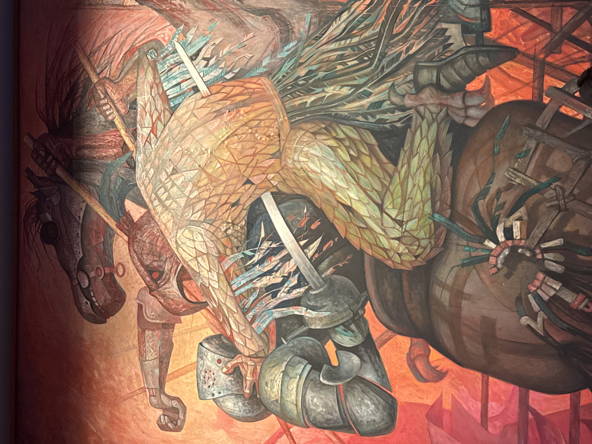 The image shows a section of a mural in Mexico City, Mexico. It depicts a violent scene with horses and riders clashing. On the left, a dark brown horse with stylized goggles and a rider in armor are visible. The rider appears to be brandishing a spear. In the center, another armored figure is entangled with a figure in indigenous dress, including a large, possibly feathered headdress. A sword is prominently displayed. On the right, another horse and rider are partially visible, also in distress and with indigenous elements. The color scheme is dominated by shades of brown, orange, and green, with a fiery red background. The style is Expressionistic and suggestive of conflict.