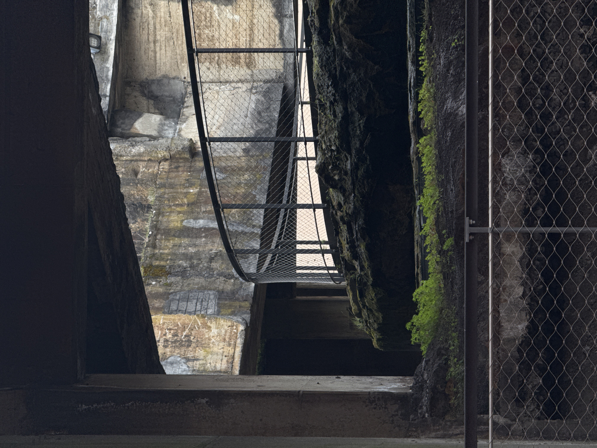 The image shows a view of a dark, possibly subterranean, concrete structure. There are concrete walls on either side and a metal mesh fence partially obscuring the view on the right. On the left, a curved metal structure runs along the concrete wall. In the center, there is a dark, cavernous opening with a rock face visible on the right side, covered in some greenery. The location is likely within the industrial area of Esch-sur-Alzette, Luxembourg, due to the presence of aged concrete structures and metal fencing.