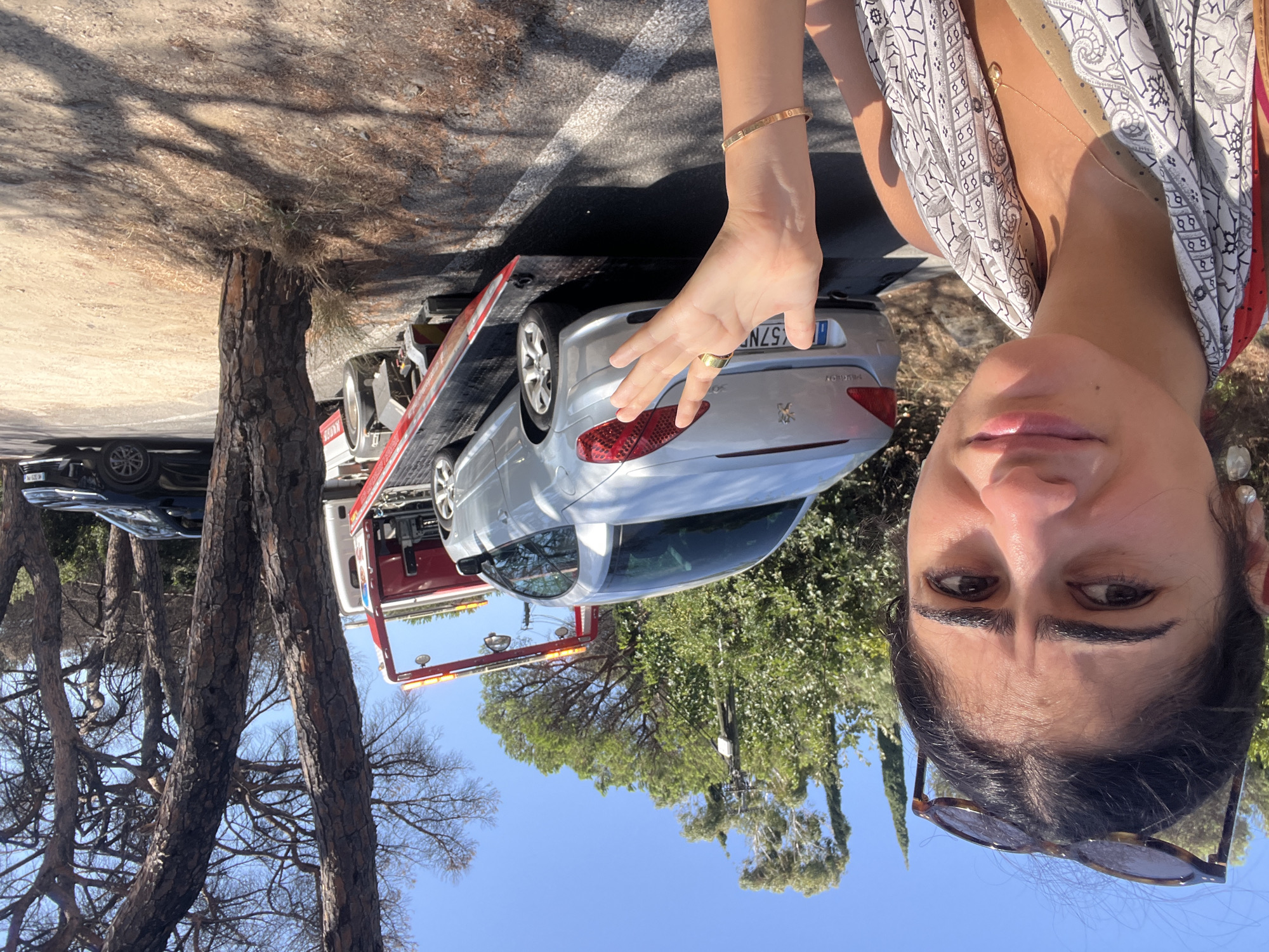 A young woman with dark hair, wearing sunglasses on her head, a patterned scarf around her neck, and a necklace, stands in the foreground of an outdoor scene. She has a worried expression and is holding up her left hand, which is adorned with rings and a bracelet. Behind her, a silver Peugeot car is loaded onto a red tow truck. To the right, a black car is parked. The background consists of trees and vegetation under a clear blue sky, indicating a sunny day. The scene appears to be in Ramatuelle, France, given the presence of a French license plate on the Peugeot. The overall impression is one of unexpected car trouble on a roadside in a rural or semi-rural area.