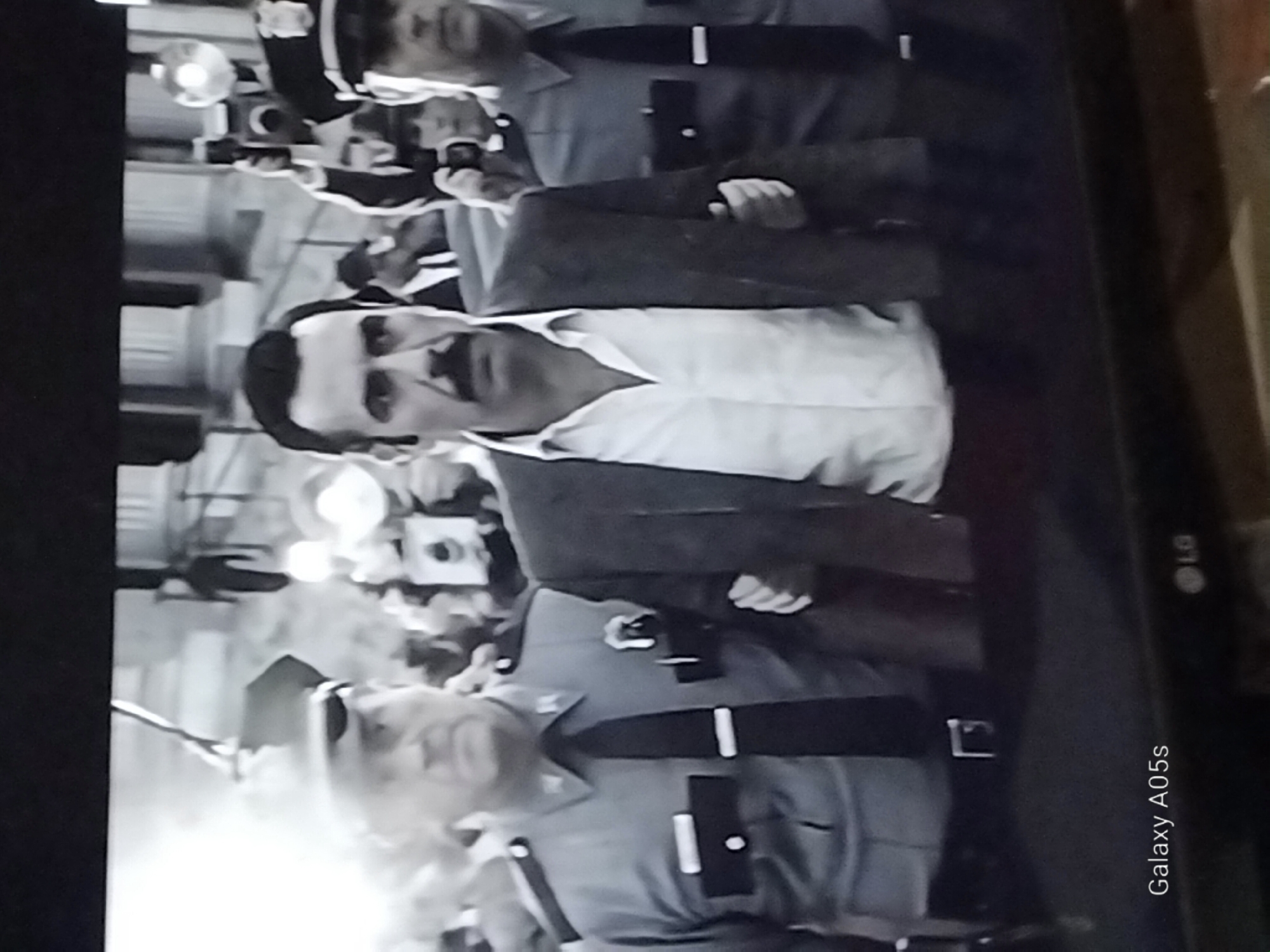 The black and white image shows a man with a prominent mustache wearing a suit jacket over a white collared shirt being escorted by two uniformed officers. The officers are wearing caps and dark ties. The man being escorted has his left arm linked with the officer on his left and his right arm linked with the officer on his right. The background appears to be an outdoor urban setting with some blurred figures and possibly buildings. There is a bright light source to the left, casting a glare on the scene. The bottom of the image displays a watermark "Galaxy A05s" and the LG logo. The image is pixelated, suggesting it was captured from a screen. There is no information available in the image to determine if this scene is located in Druzhba, Russia.