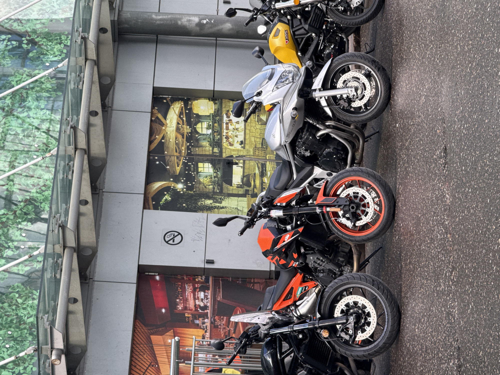 A row of four parked motorcycles is captured in a low-angle, medium shot on a wet asphalt surface. The motorcycles are positioned side-by-side, facing left. The leftmost motorcycle is predominantly black, followed by an orange and black KTM, a silver Suzuki, and a yellow motorcycle with black accents on the far right. The motorcycles have visible engines, wheels with disc brakes, and handlebars. Behind the motorcycles, a modern building facade features large graphical panels depicting interior scenes of a bar and a restaurant with warm lighting and chandeliers. Above these panels, a glass canopy with exposed metal supports is visible, reflecting green foliage from an overhead installation. The overall environment suggests an urban setting. There are no people visible in the image. The weather appears to be overcast, indicated by the wet ground. A "no smoking" sign is present on a light-colored panel between the first two graphical panels.