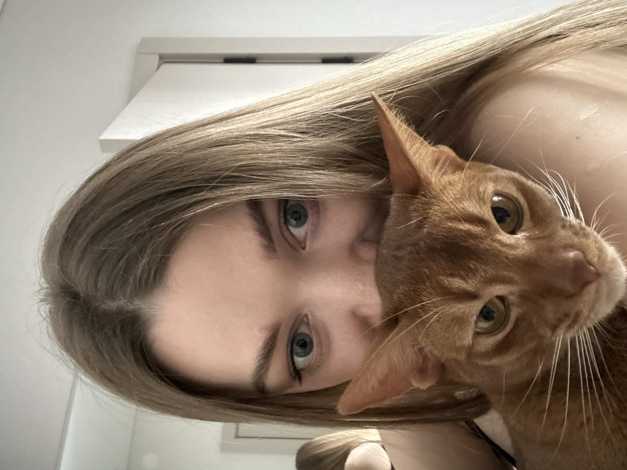 A close-up, portrait-style photograph features a young woman and an orange tabby cat. The woman has long, straight blonde or light brown hair that frames her face. Her visible left eye (from her perspective) has a striking blue-green iris, and her gaze is directed towards the viewer. Her fair skin and visible shoulder suggest she is bare-shouldered or wearing light clothing.
Cuddled intimately beside her, with its head resting near her face and shoulder, is an orange or ginger cat. The cat's large, expressive eyes, which appear amber or light green, are also looking towards the camera. Its prominent white whiskers extend outwards, and its fur is a vibrant reddish-orange hue.
The image conveys a sense of closeness and affection between the two subjects. The lighting is soft and diffused, suggesting an indoor setting, possibly a home. A plain white wall and the edge of a white doorframe are visible in the background, hinting at a minimalist interior. There are no specific visual cues for time of day, weather, or visible text. The provided location context for the image is Kazan, Russia.
