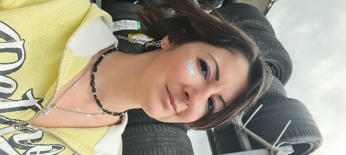 An adult female is depicted in a medium close-up shot, positioned outdoors in the city of Mate Asher Regional Council, Israel. She has short, dark brown hair and is looking towards the viewer with a subtle smile. Her eyes feature purple eyeliner on the upper lids, and silver glitter is applied to her cheekbones beneath both eyes. She has facial piercings, including a septum ring and a nostril stud. The female wears a yellow hooded sweatshirt with a partially open zipper, revealing a dark pattern on the garment's front. Her visible left ear displays multiple earrings, including hoops and a dangling charm with a crescent moon and chain. She wears two necklaces: a black beaded choker with silver accents and a longer silver chain with a pendant. Silver glitter is also visible on her upper chest. In the background, multiple black vehicle tires are stacked on metal racks. The sky above is overcast and light grey. A green and white label is partially visible behind her left shoulder.