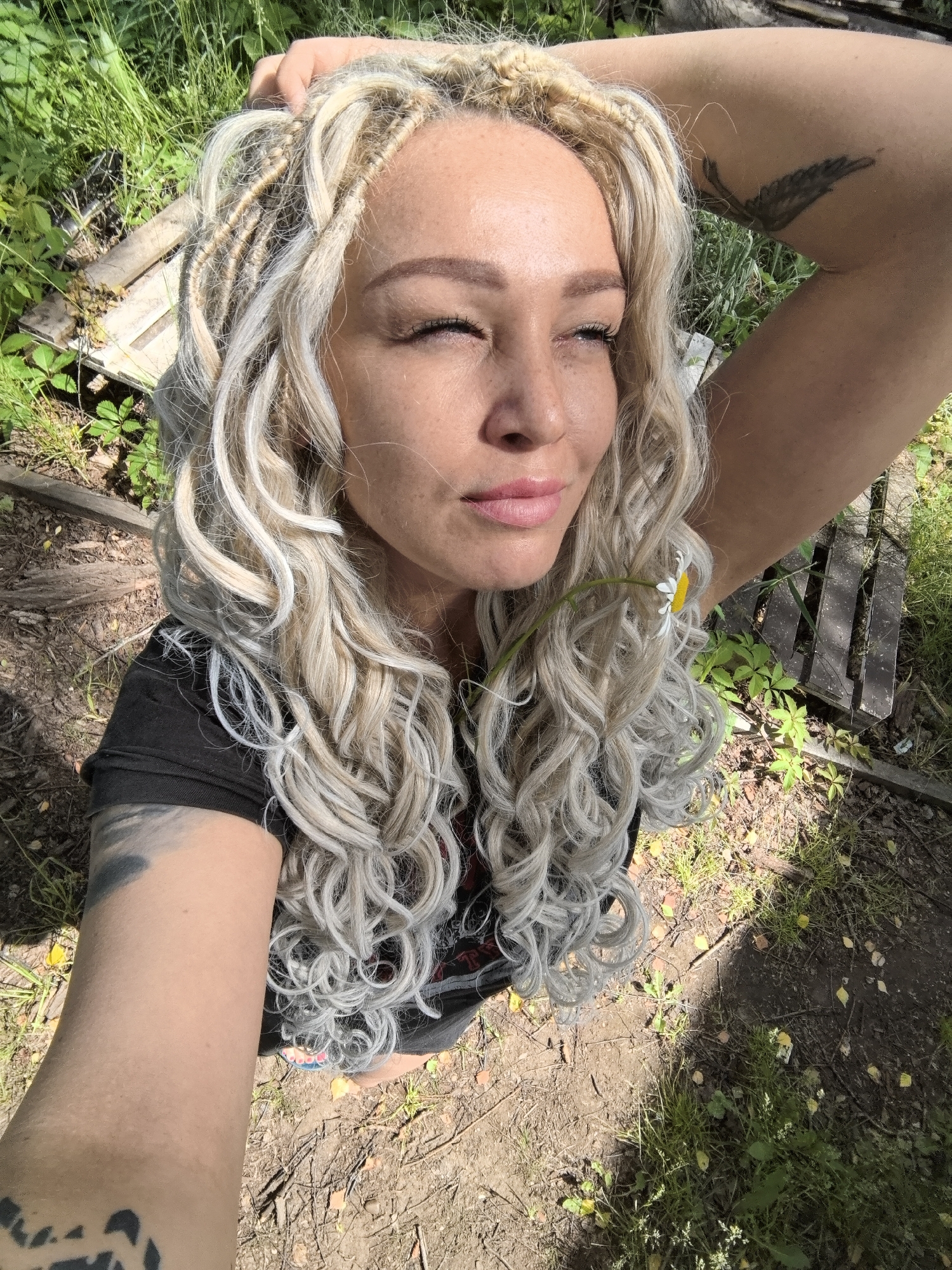 A medium shot photograph captures a woman with blonde dreadlocks from a high angle. She is outdoors, with sunlight casting shadows. The woman is wearing a black t-shirt and has tattoos on her arms. Her eyes are closed, and she is touching her head with one hand. A daisy is tucked into her hair. The background consists of grass, dirt, and wooden planks, suggesting a rural or park-like setting. The location is identified as Noviy, Russia.