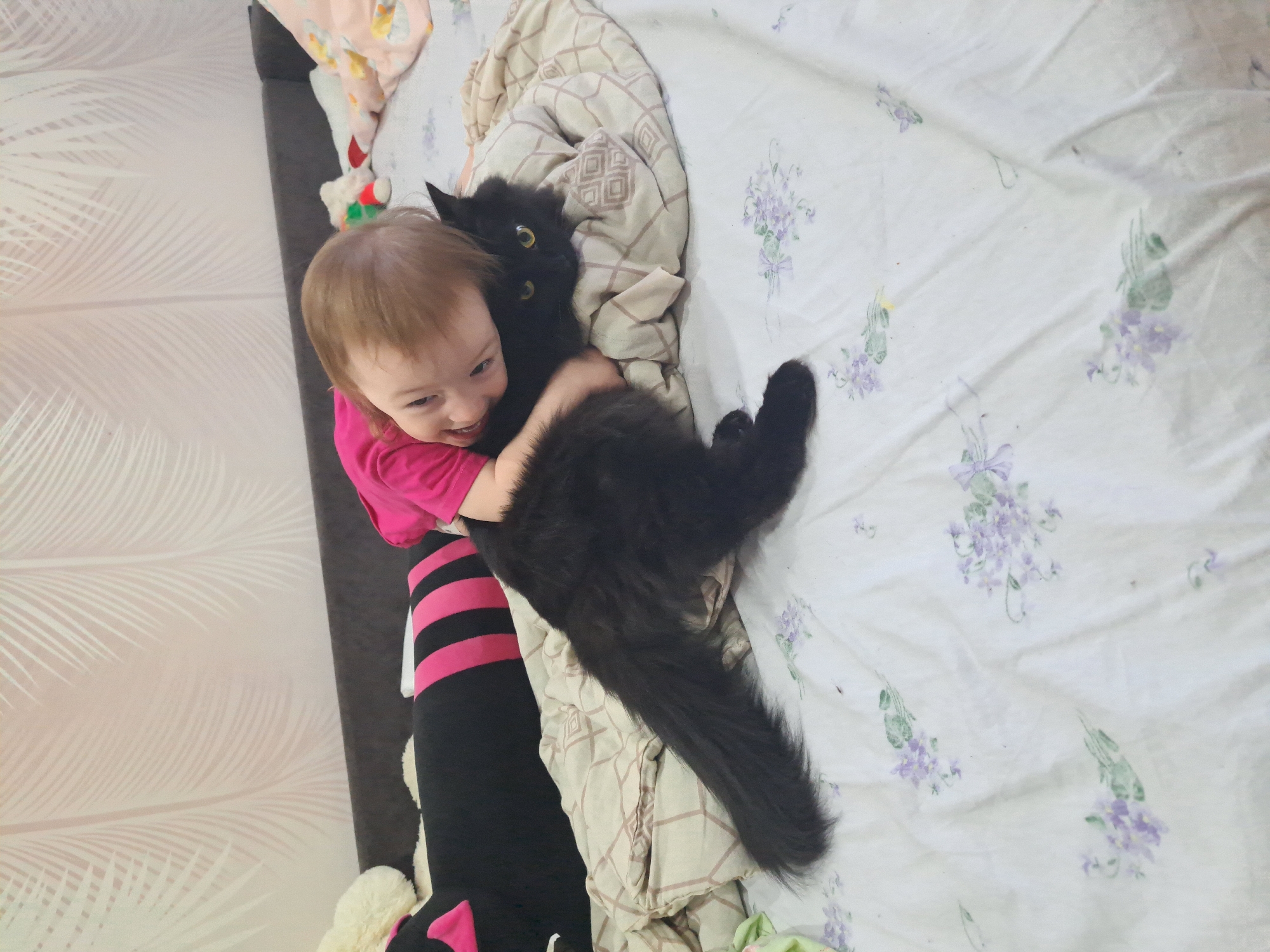A child embraces a black cat on a bed in Chaikovsky, Russia. The child, with light brown hair, wears a pink shirt and striped leggings. The black cat is on a white floral-patterned bedsheet with a beige geometric-patterned blanket. A stuffed animal is visible near the headboard. The wall has a light feather-like pattern.