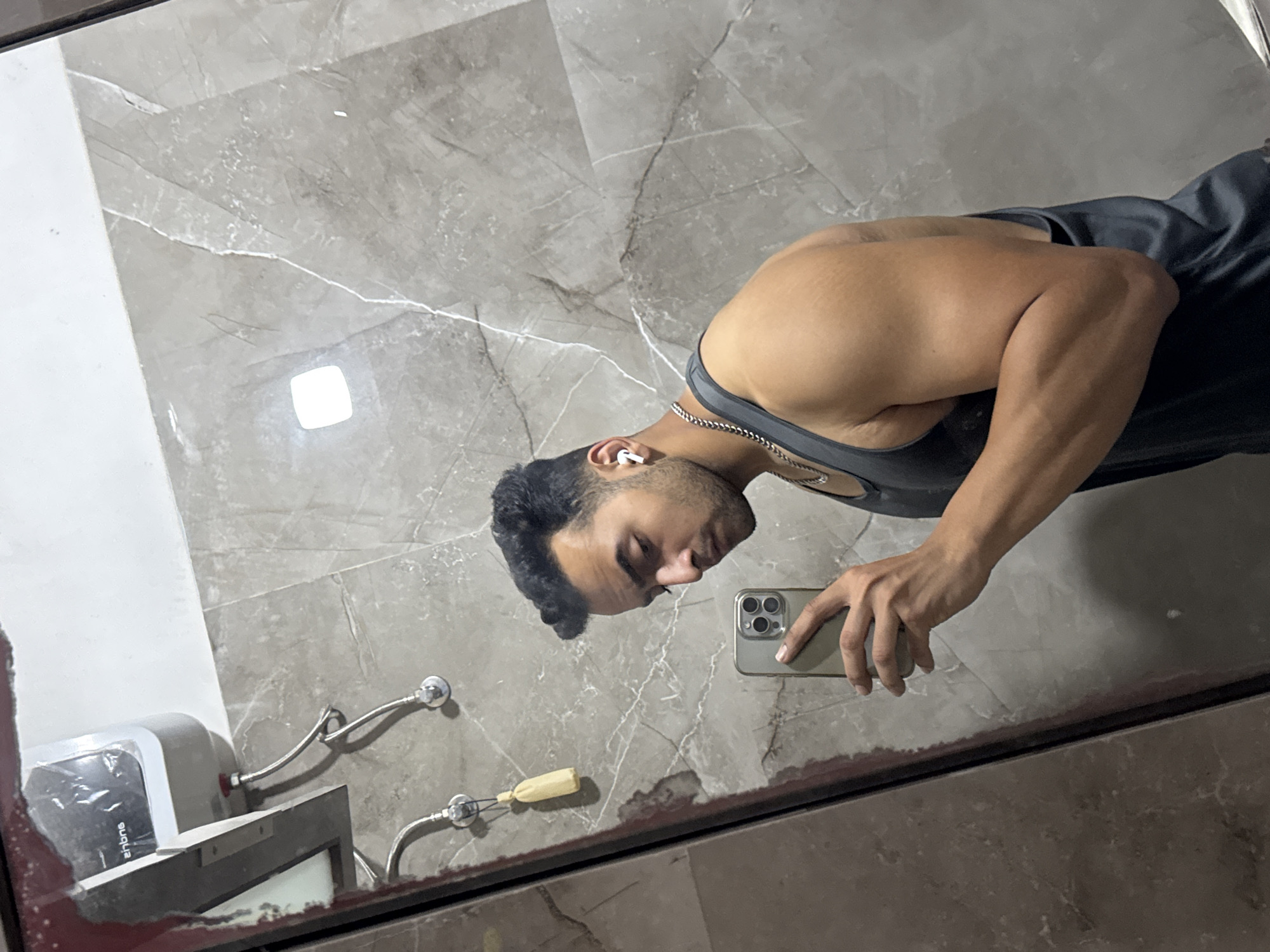 A shirtless man with dark hair and a chain around his neck takes a selfie in a bathroom. He is wearing earbuds and holding a smartphone with three cameras. The reflection shows a sink, faucet, and a hot water dispenser mounted on a marble-like tile wall. The location is likely in India, specifically Kolkata, as inferred from the "Antone" brand on the water dispenser, which is a brand common in the Kolkata market.