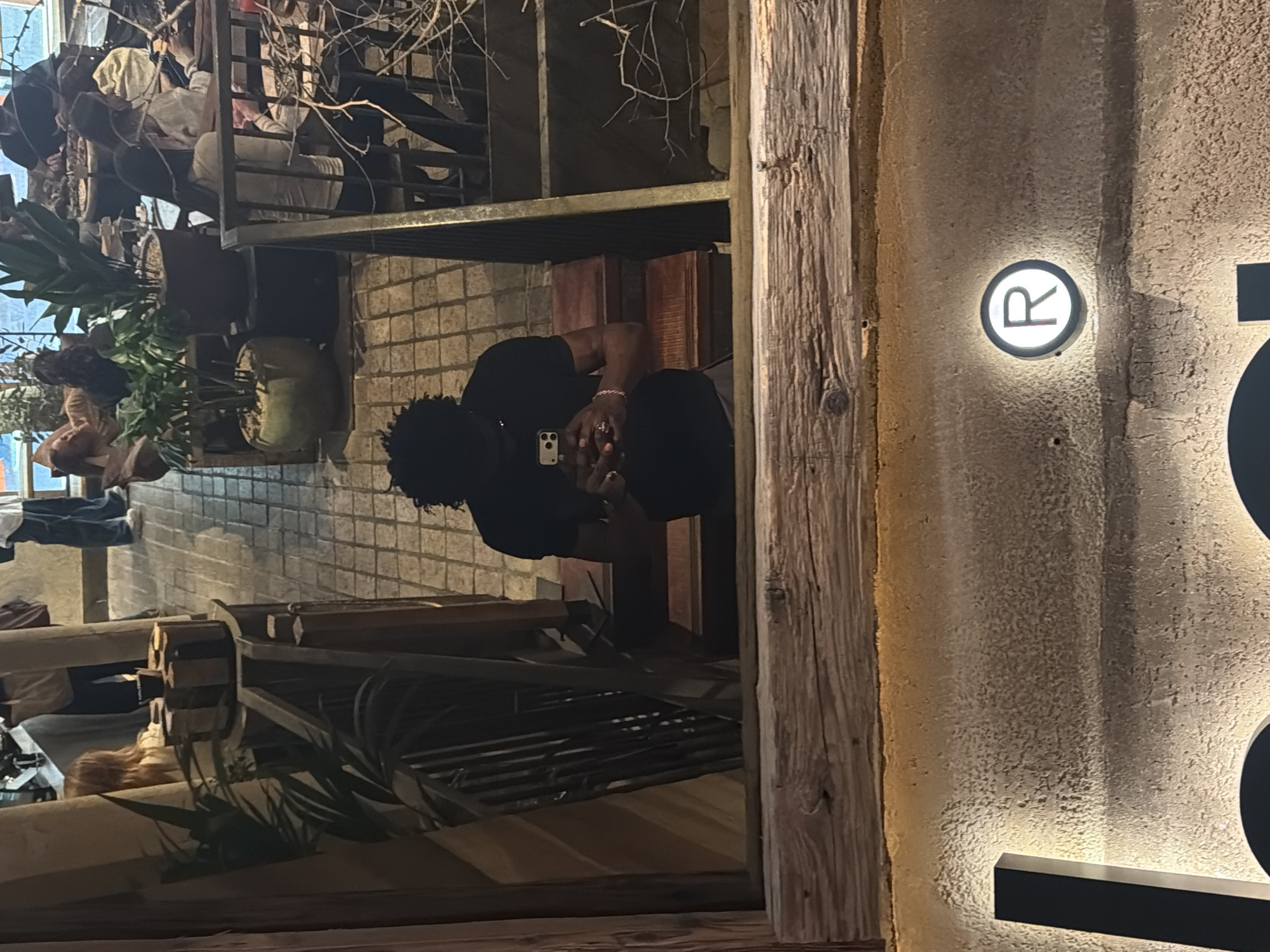 The image depicts an individual seated at a table within a restaurant or bar setting. The person, possibly male, is wearing dark clothing and appears to be engaged with a mobile phone. The setting includes shelves, plants, brickwork, and an industrial aesthetic. The image features a lit logo with a letter 'R', suggesting the venue name. The exterior wall texture of the building is coarse, likely concrete.
Although an exact location cannot be determined with certainty from the image, the style of the restaurant is reminiscent of urban dining establishments in London. The combination of industrial elements, modern lighting, and fashionable clientele aligns with upscale dining options found across various central London locations. It is not possible to definitively identify the specific establishment or area of London without additional external information or features like a street sign.