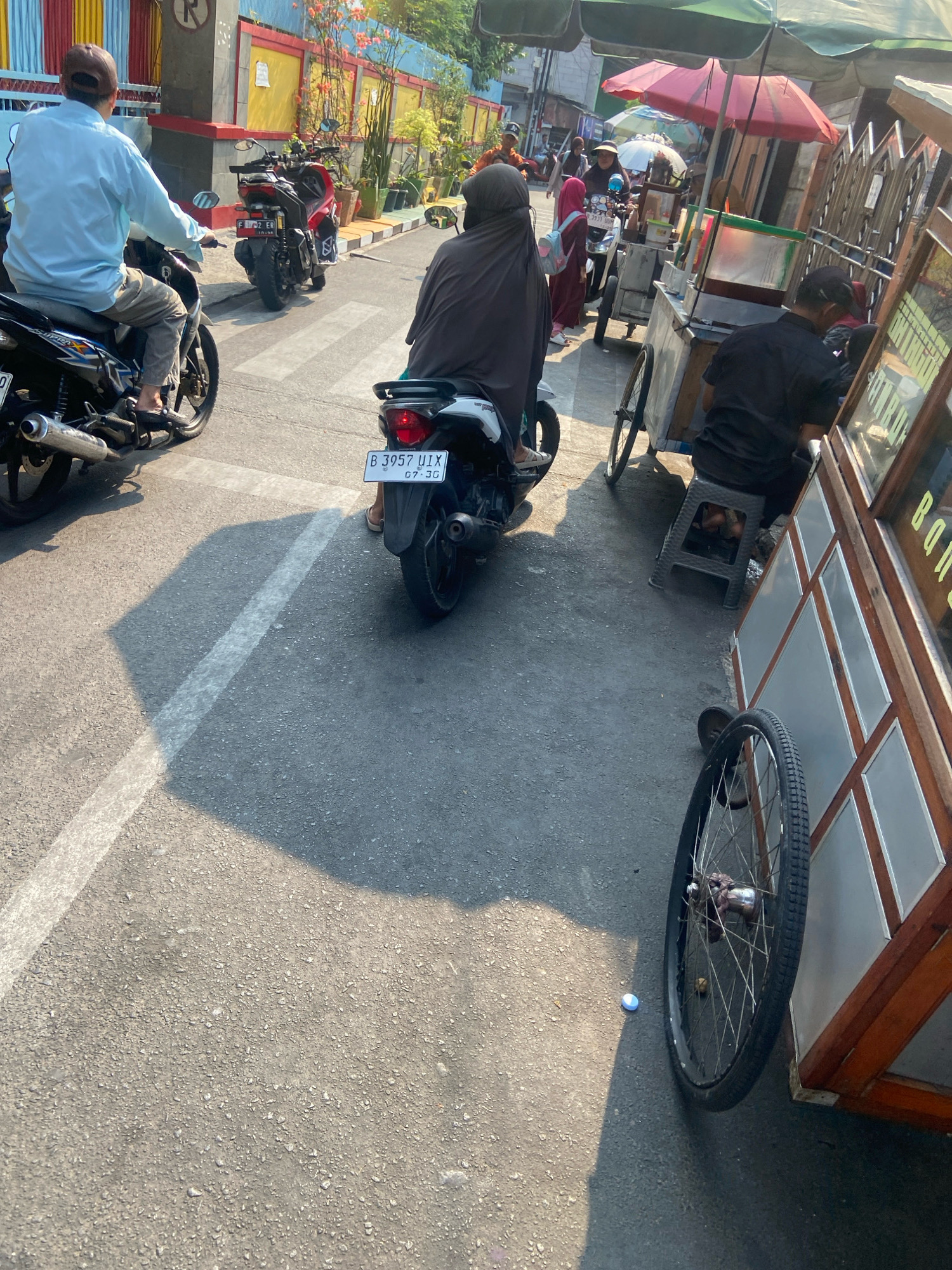 This image depicts a street scene in North Jakarta, Indonesia, likely a local marketplace or roadside. The foreground is dominated by a paved road with a white dashed line, casting long shadows from the midday sun. On the left, a person wearing a light blue long-sleeved shirt and grey pants rides a black motorcycle. Behind them, another motorcycle, this one red, is parked. In the center, a person wearing a black hijab rides a scooter. The license plate reads "B 3957 UIX". To the right, a food cart with a wooden frame and glass panels is visible, with a person in a black shirt sitting on a plastic stool nearby. Another person, partially visible, is also present near the cart. In the background, a colorful building with red and yellow accents lines the street, with potted plants adding greenery. Several other people and vehicles are scattered throughout the scene, suggesting a busy, everyday environment. The overall impression is that of a typical urban street in Indonesia, with a focus on pedestrian and motorcycle traffic, small businesses, and daily life.