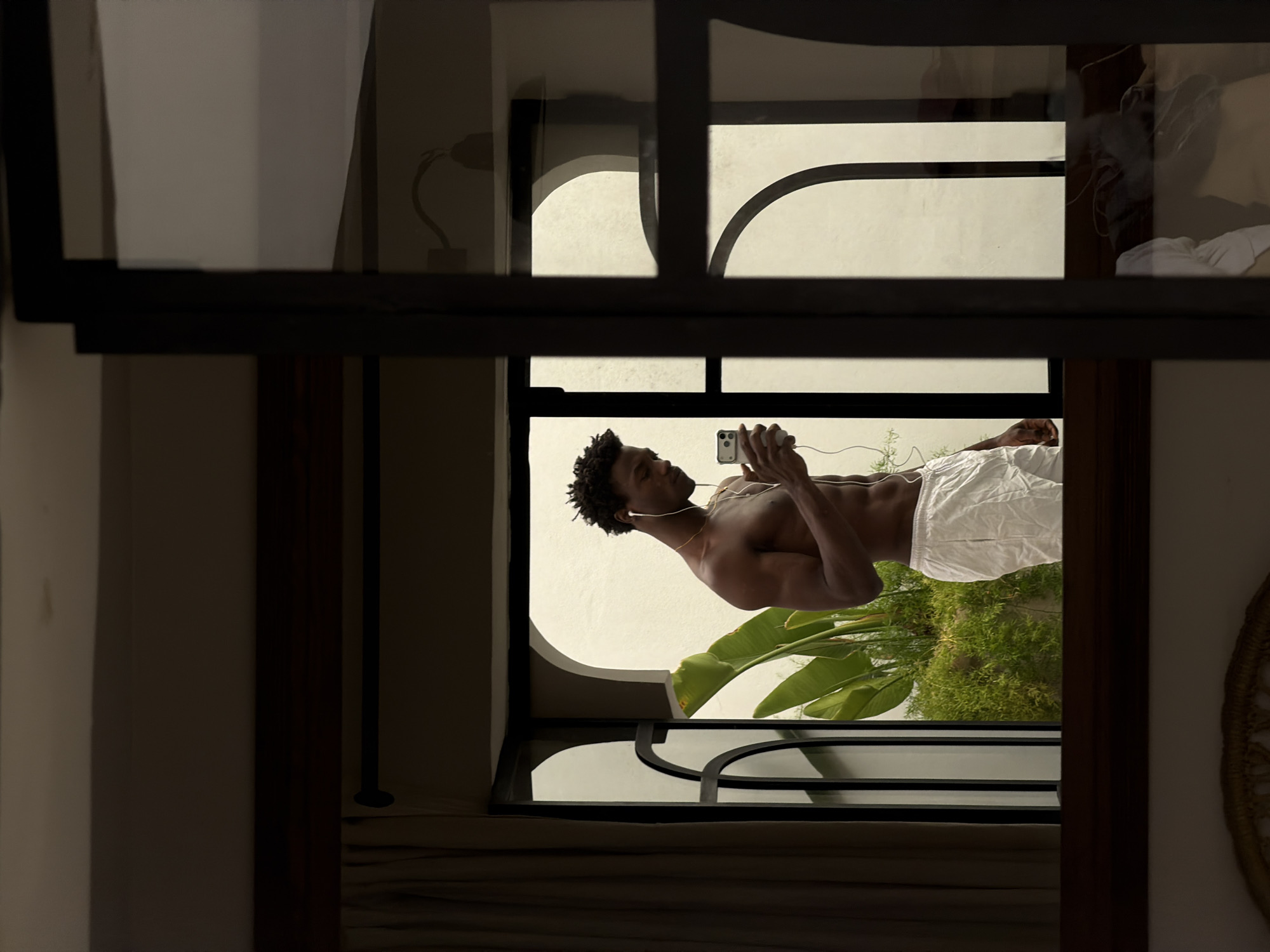 The image depicts a shirtless young Black man standing by an arched window, holding a smartphone in his right hand and wearing white earbuds. He is looking down at the phone, with his left hand near his chest. He is wearing white shorts. Lush green foliage is visible through the window. The scene appears to be in an indoor setting with a dark-framed window or doorway in the foreground, suggesting a reflection. The architectural style of the window, with its rounded arches, and the presence of exotic plants are characteristic of the city of Marrakesh, Morocco.