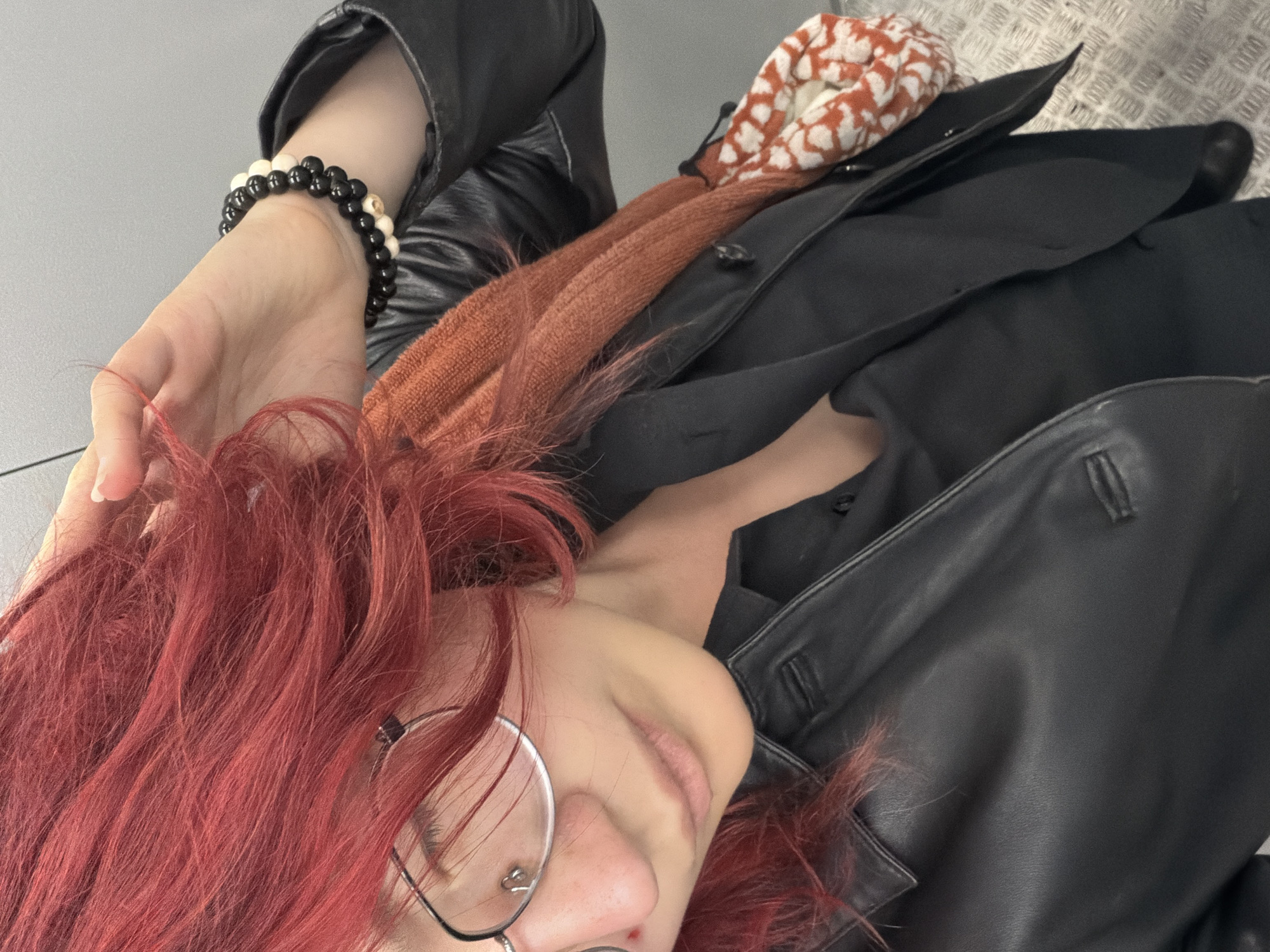 The image shows a close-up of a young person with vibrant red hair, wearing glasses with a silver frame and a nose ring. They are wearing a black collared shirt and a black leather jacket. A burnt orange scarf is draped around their neck, and a small bag with a white and orange patterned design hangs from their shoulder. They are also wearing a bracelet made of black and white beads on their wrist. The background appears to be a neutral gray wall. The lighting suggests an indoor setting. The person's gaze is directed downwards, and they are touching their hair with their right hand. The flooring visible at the bottom of the image is a metal grate pattern. There is no identifiable information to determine the location in the city of Chelyabinsk, Russia.