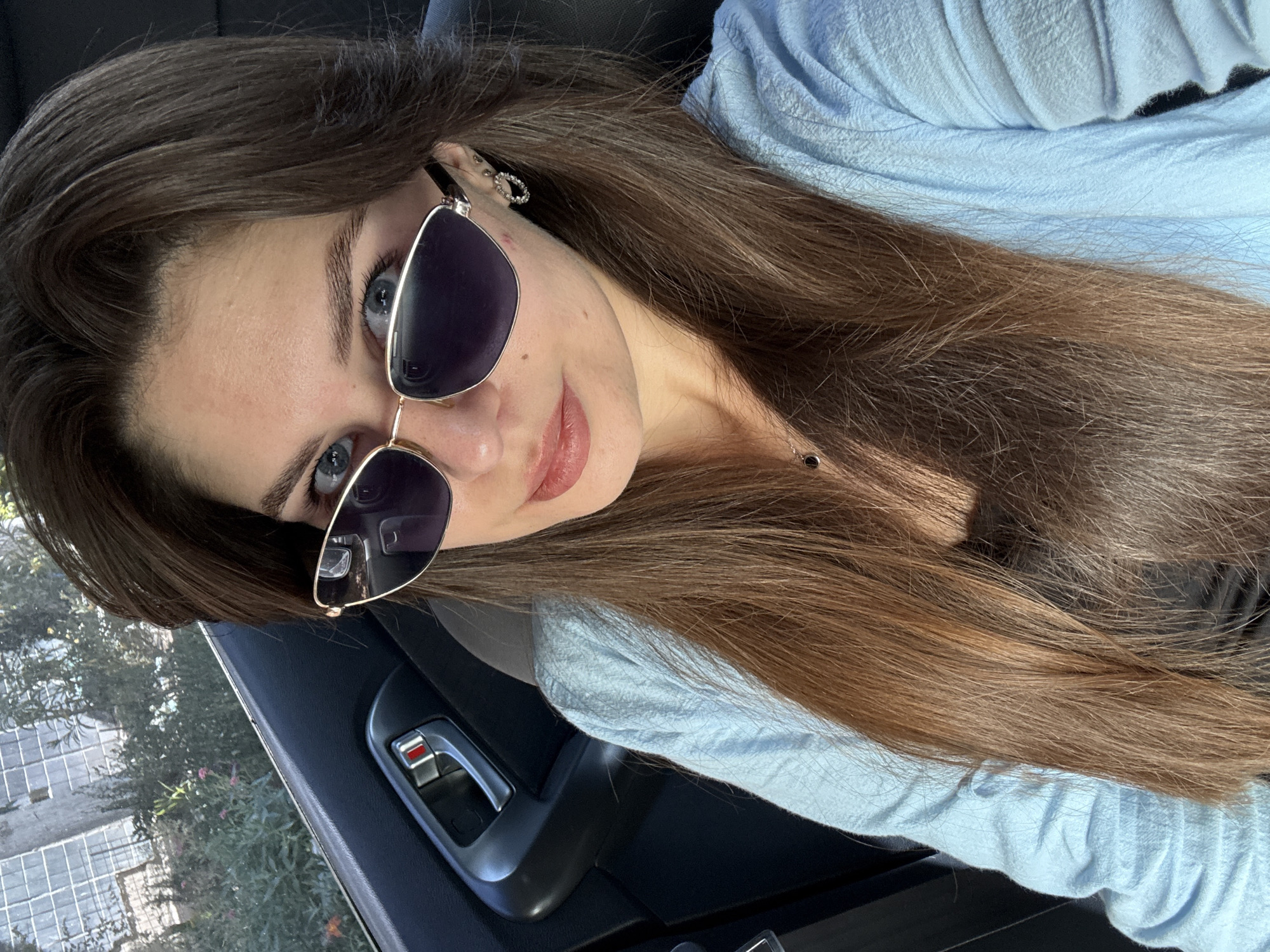 The image is a close-up, selfie-style photograph of a young woman with long, brown hair and blue eyes. She is wearing large, dark-lensed sunglasses with gold frames and a light blue top. She has a small stud earring in her visible ear. Her skin appears clear, with a light dusting of freckles across her nose. Her lips are slightly parted, and she has a subtle smile. The lighting suggests it is daytime, possibly outdoors or in a well-lit car interior, as the sunlight catches the highlights in her hair.
The background is partially visible, showing the dark interior of a car, including a door handle and window. Through the window, trees and a building with a grid-like structure can be seen, indicating an urban or suburban setting. The orientation of the image is tilted, adding a dynamic perspective to the shot. The woman's gaze is directed towards the camera, conveying a casual and confident expression. There is no visible text in the image.