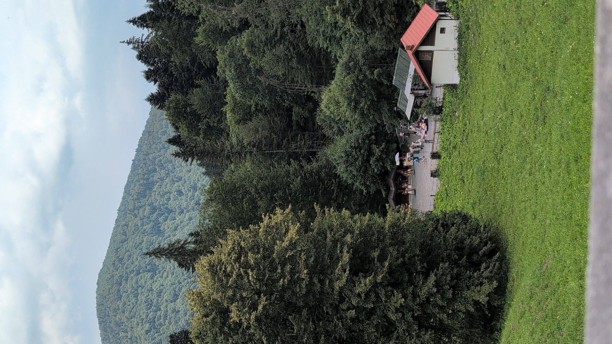 The image depicts a natural scene in Sinaia, Romania, featuring a lush, forested mountain slope in the background under a cloudy sky. In the mid-ground, there is a clearing with a small village-like settlement. This settlement includes several structures, including two covered stalls or kiosks, and a small white building with a red roof, suggesting a recreational or tourist area. A group of people, appearing to be tourists or visitors, are gathered in the clearing, some browsing at stalls, while others are standing or walking. The foreground is dominated by a gently sloping green meadow. The overall atmosphere is that of a peaceful, outdoor environment, likely a mountain resort or a park.