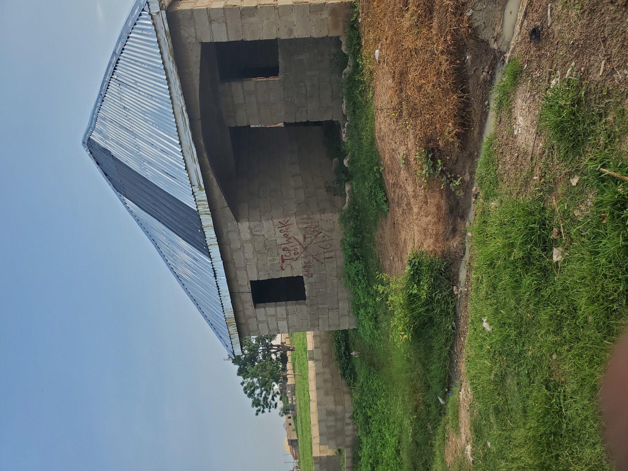 The image depicts an unfinished cinder block building with a corrugated metal roof. Red spray paint graffiti reading "STOP WORK" and other illegible text is visible on the side of the building. In the foreground, there is overgrown grass and a small ditch with murky water. In the background, other unfinished structures and trees are visible under a clear blue sky. There is no indication of the city of Winters, United States of America. A chicken is visible walking in the grass in the mid-ground.