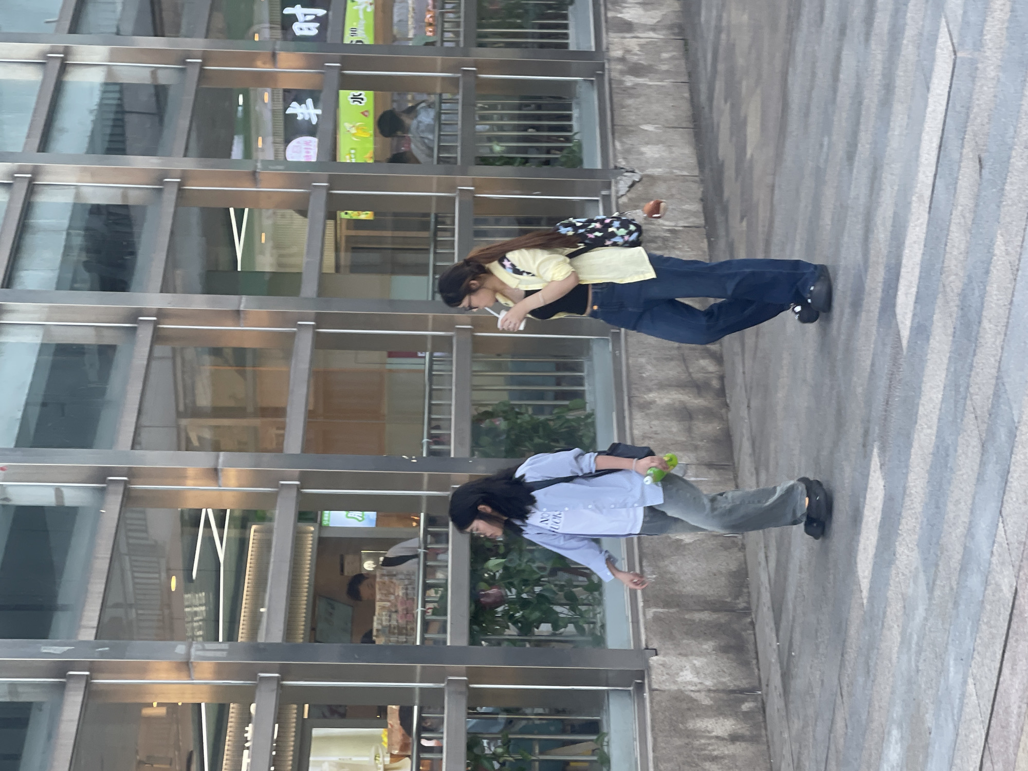 The photograph depicts two young women walking side-by-side on a paved walkway in an urban setting. A young woman on the left wears a light blue shirt and carries a green bottle. A black bag hangs over her shoulder. The second woman, positioned to the right, is drinking from a cup and has a multi-colored backpack on. She wears a yellow jacket, a black top, and blue jeans. They both face the same direction and appear to be talking. A building with large glass windows and metal framework is behind them. The location is in the High-tech Zone of Chongqing, China. The background includes details of shops and interiors visible through the building's windows.