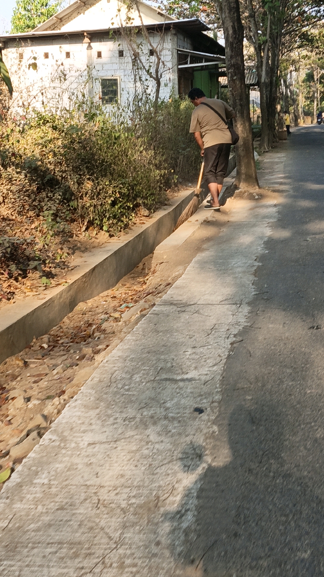 A person, identified as a man, is sweeping a concrete drainage ditch alongside a paved road. He is wearing a light brown t-shirt, dark shorts, and black shoes. A dark bag is slung across his right shoulder. The man is facing away from the camera, towards the right side of the image, and is actively using a broom. The drainage ditch is filled with dirt, leaves, and debris. To the left of the ditch is a dense patch of green bushes, and behind the bushes is a white, low-rise building with a metal roof and several small square windows. Several trees with visible trunks line the right side of the road, casting shadows on the pavement. Further down the road, another person is visible in the distance, wearing yellow. The scene is outdoors, with sunlight illuminating the area. The overall impression is of a street cleaning activity in a residential or semi-rural area. The location is specified as Kuwasen, Indonesia.