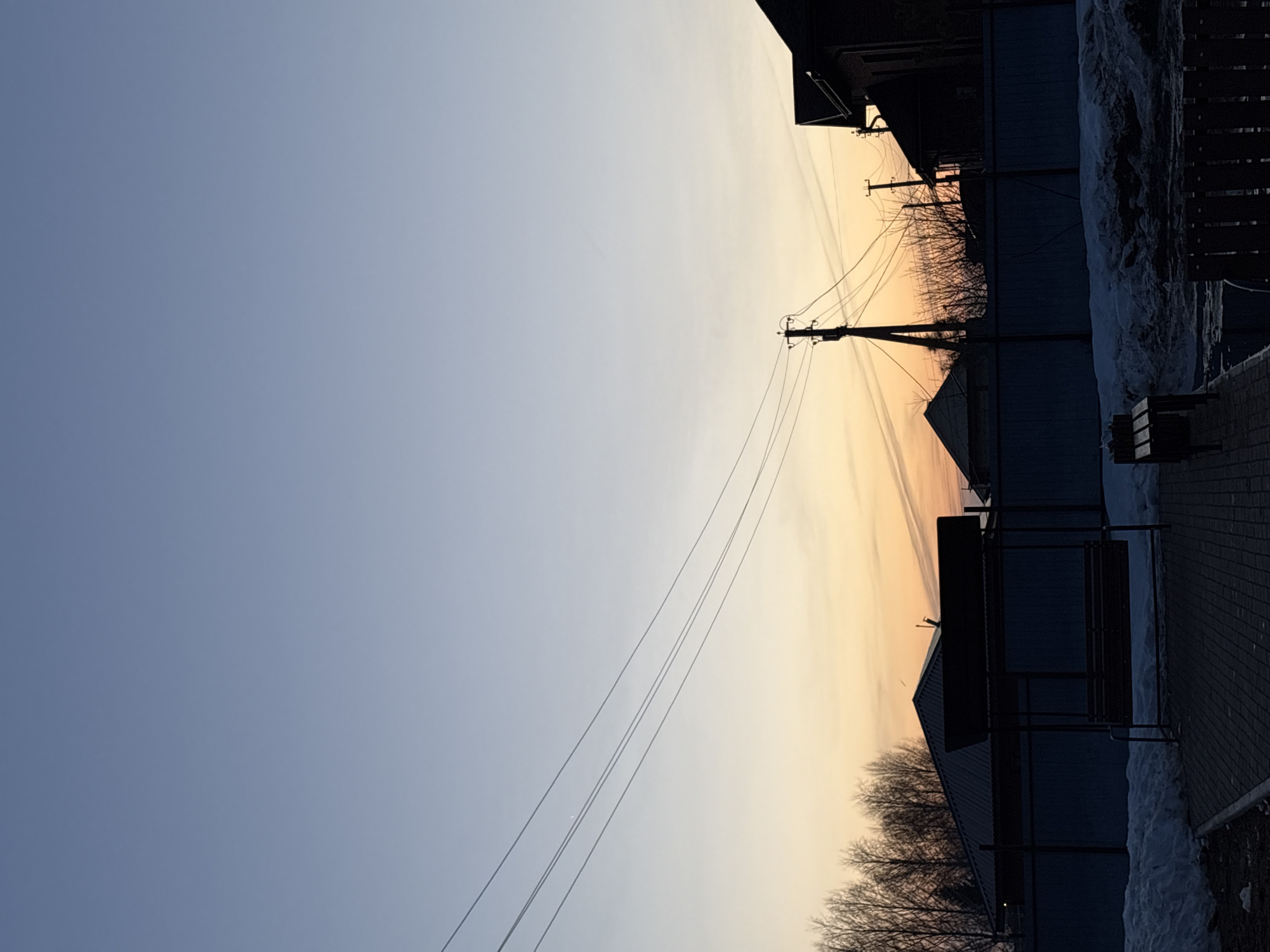 The image shows an urban scene in Прикамский, Russia, with a view towards the horizon during sunset or sunrise. The sky transitions from a pale blue to a warm orange glow. Power lines extend diagonally across the sky from a utility pole. In the foreground, there are buildings and bare trees silhouetted against the sky. A brick walkway borders the scene, with patches of snow visible along the edge. A bus stop structure is also visible along the side of the walkway.
