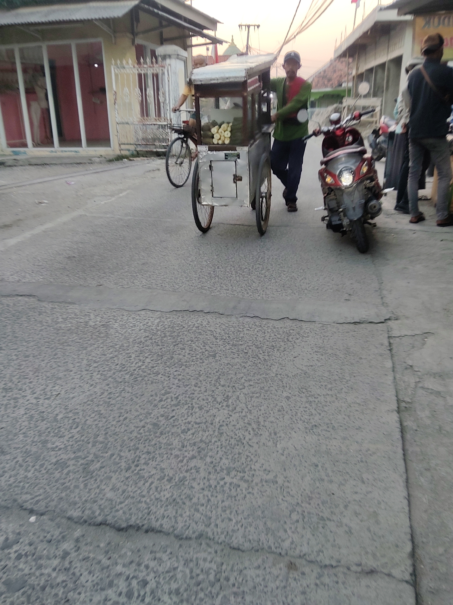 In Mekarmukti, Indonesia, a street scene unfolds at dusk with a vendor pushing a food cart. The cart, made of metal and glass, displays fruits, possibly durian, in its lower section. A man, the vendor, wearing a green shirt and a red scarf, stands beside the cart, his hand resting on its structure. To the right of the cart, a red motorcycle is parked, its headlights illuminated. Further to the right, two individuals are visible, one in a dark shirt and pants with a brown cap, and another in a lighter colored shirt and dark pants. In the background, a building with a corrugated roof and large windows is situated, hinting at a commercial area. The street is paved with asphalt, showing signs of wear and tear, with a visible crack running across it. The sky is a soft orange hue, indicative of the twilight hour.