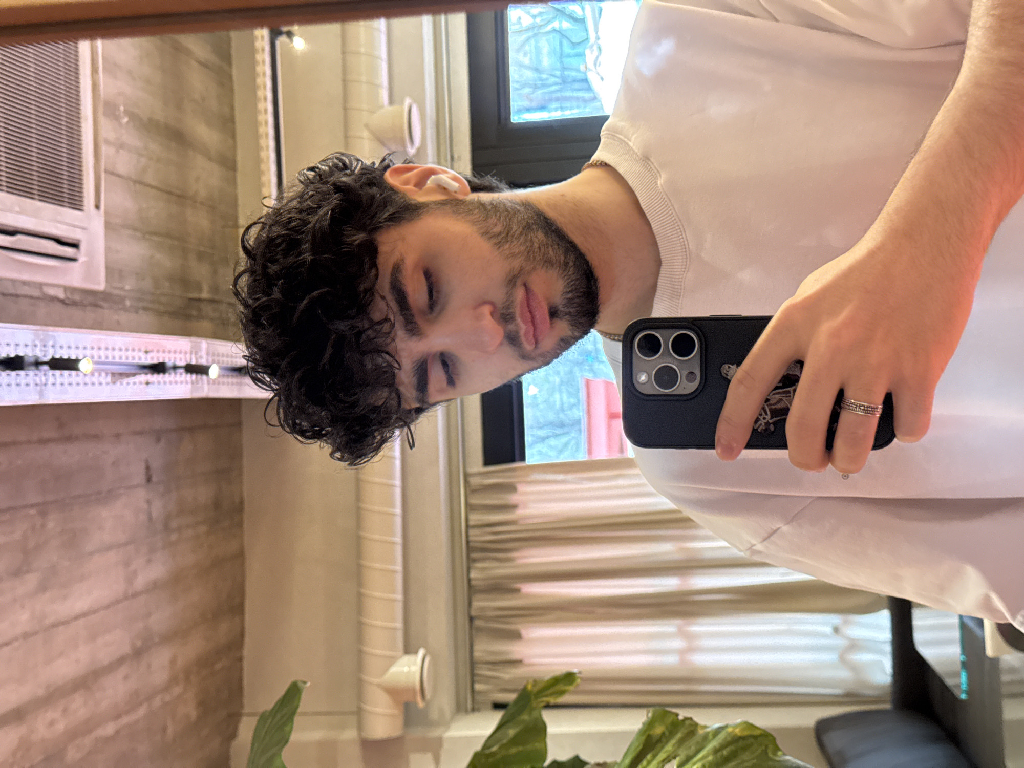 A man is taking a selfie in a mirror. He has dark curly hair, a beard, and is wearing a white t-shirt. He is holding a black smartphone with a triple-lens camera. He is wearing an earpiece in his right ear and has a ring on his finger. The background shows a room with light-colored walls, a window, an air conditioning unit, and what appears to be a plant. There is no clear indication that this image was taken in Yerevan, Armenia.