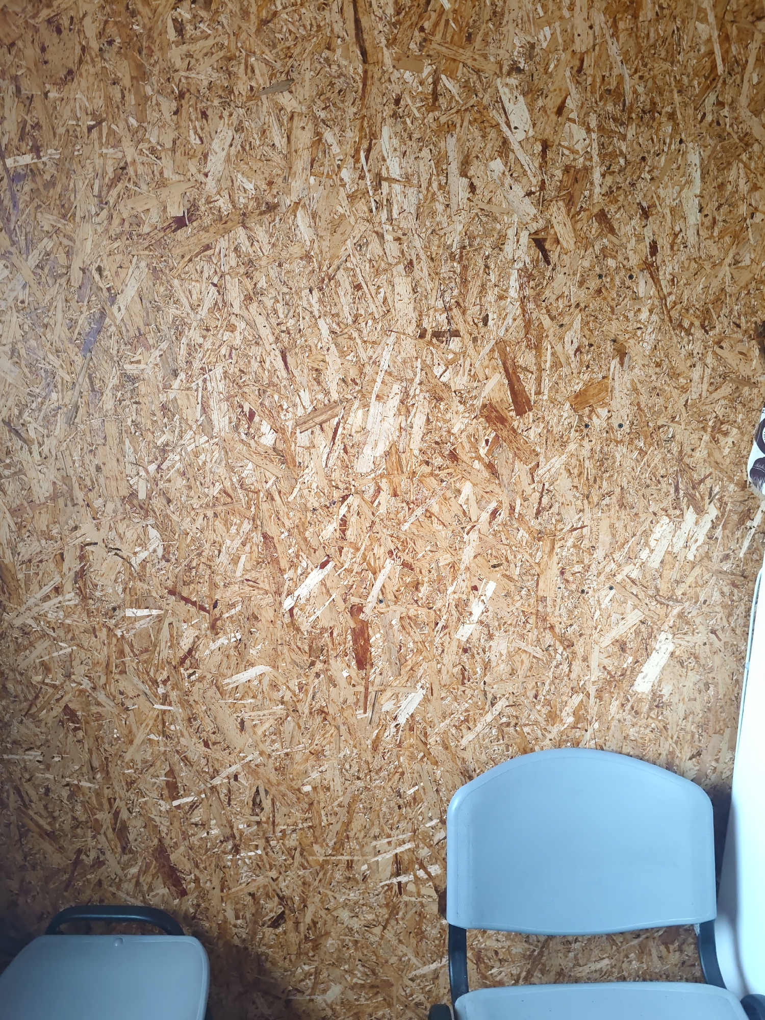 The image shows the interior of a building with walls made of oriented strand board (OSB). In the foreground, two light blue chairs are partially visible. The chair on the right has a distinct curved back. The chair on the left appears to be a foldable stool with a dark handle. A portion of a white object, possibly a whiteboard or partition, is visible on the far right edge of the image. The scene is likely a room in a school, community center, or similar facility in the village of Platoschino, Russia, given the functional and somewhat sparse furnishings. The OSB walls suggest a utilitarian construction. There are no people present in the image.