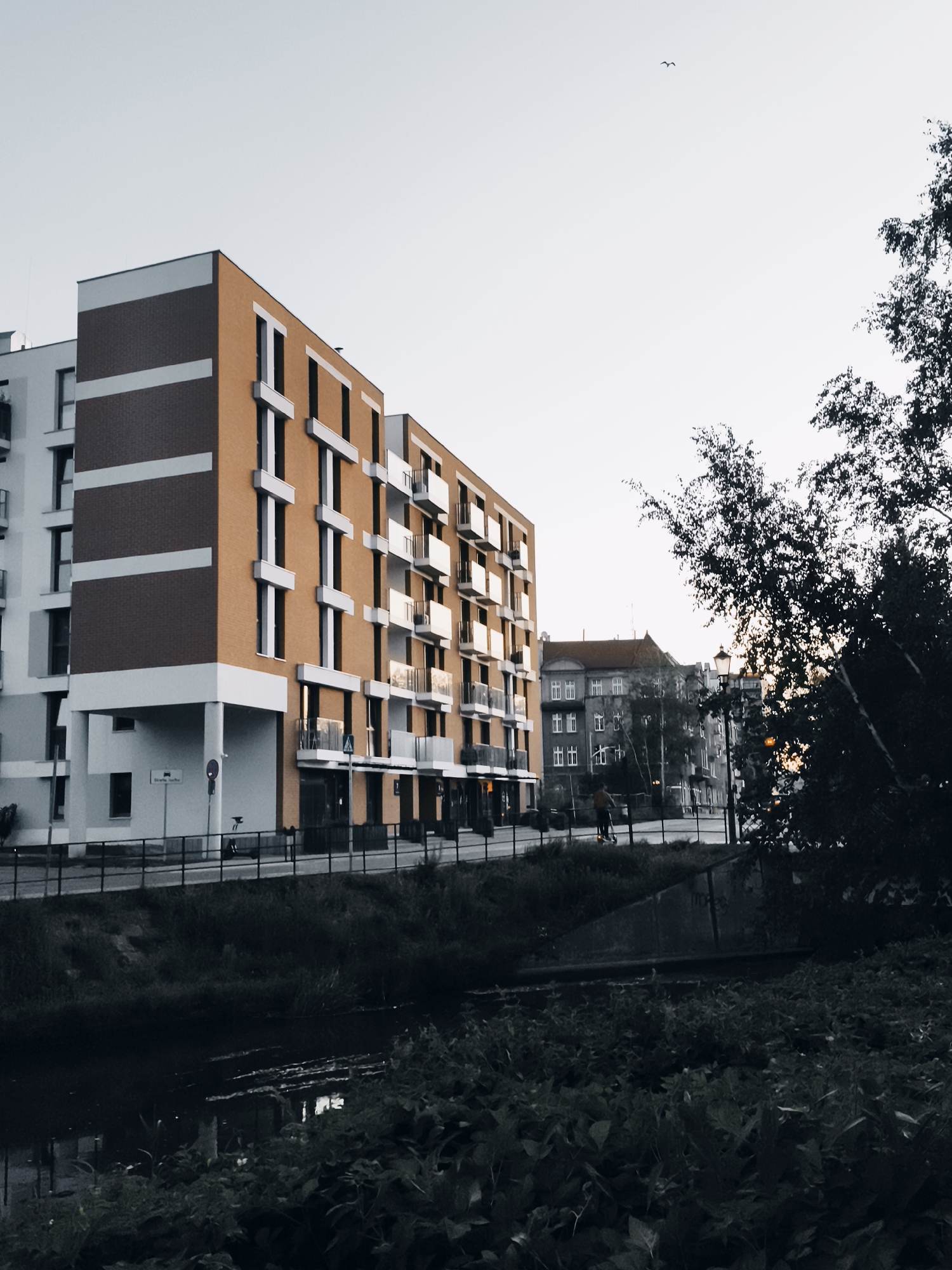 The image captures a modern apartment building with orange-brown brick facade and white accents, located in Gdańsk, Poland. The building features multiple balconies with glass railings and large windows. Adjacent to it is another, older building with a red-tiled roof. In the foreground, a dark body of water, likely a canal or river, reflects the sky and surrounding vegetation. Lush greenery lines the bank of the water. A person on a scooter is visible on the sidewalk in front of the buildings. The sky is overcast, suggesting it might be early morning or late afternoon. The overall scene depicts an urban environment with a blend of contemporary and older architecture, bordered by natural elements.