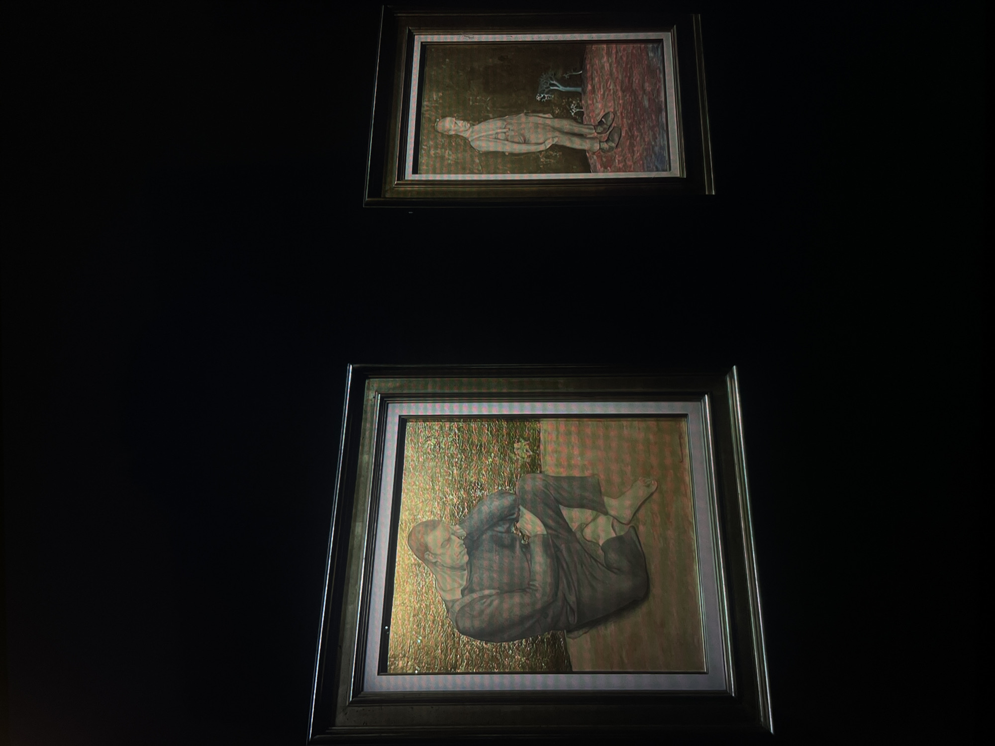 Two framed paintings hang on a dark wall. The upper painting depicts a man lying prone in a landscape. The lower painting depicts a man curled in a fetal position. The location is Unknown, Unknown.