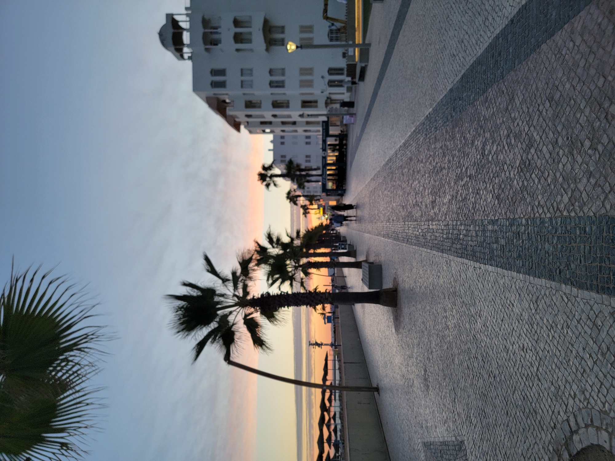 An outdoor, eye-level, full shot captures a seaside promenade at dusk in Quarteira, Portugal. The paved walkway, made of grey cobblestones, leads towards a low-rise white building adorned with balconies. Palm trees line the left side of the promenade, casting long shadows on the pavement. In the distance, beach umbrellas are visible along the shore, and the sky displays the soft hues of sunset with streaks of pink and orange. A few people are seen walking along the promenade. The scene is tranquil, with the light fading as evening approaches.
