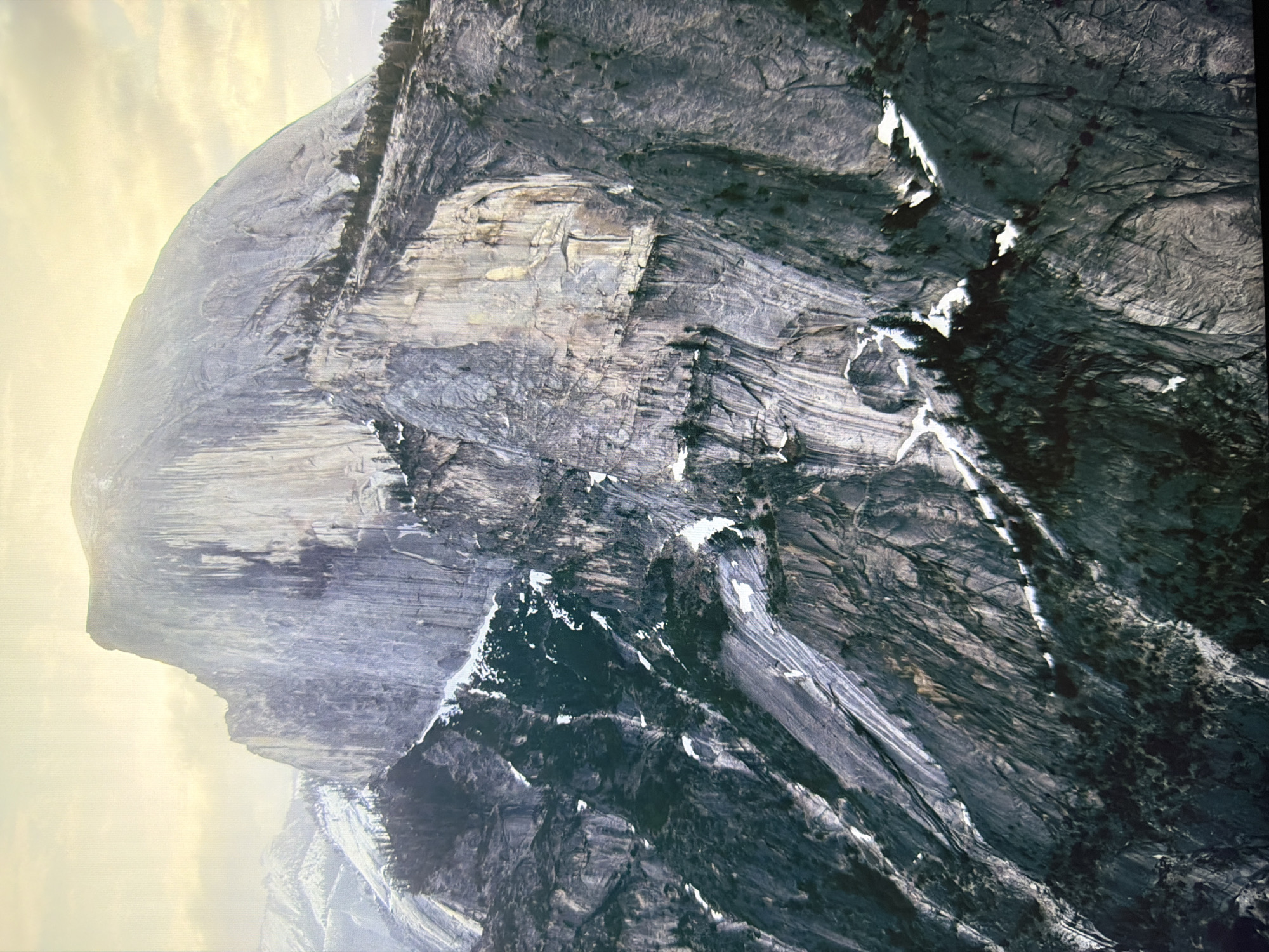This is an aerial photograph of Half Dome in Yosemite National Park, California, USA. The image captures the iconic granite dome from a high vantage point, with the sun illuminating its sheer face. The slopes leading up to the dome are covered in a mix of dark green coniferous trees and patches of snow. The sky above is bright and hazy with some clouds. There are no people or identifiable man-made structures visible in the image, nor is there any connection to the city of Kyiv, Ukraine.