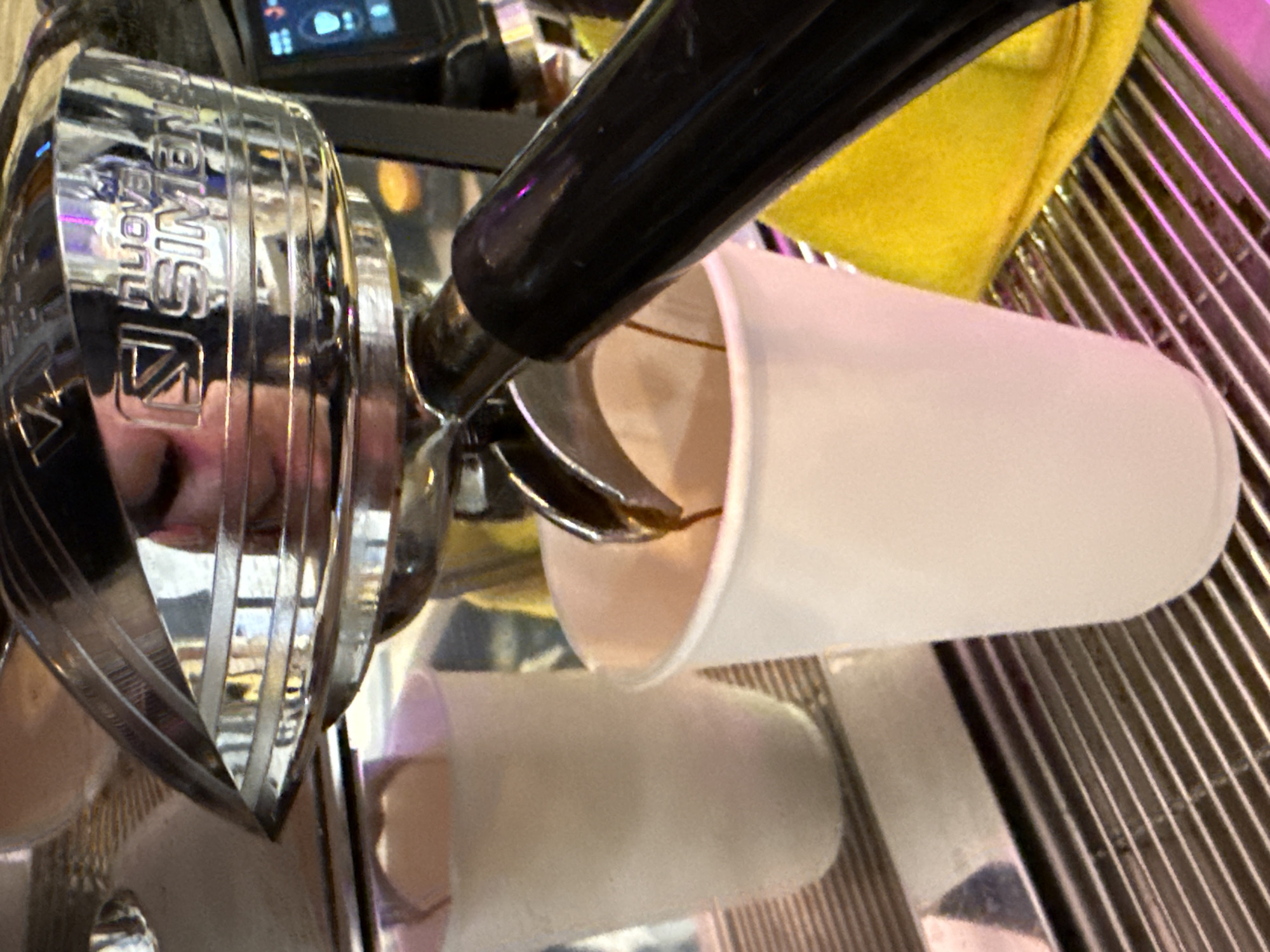This is a close-up, eye-level shot of an espresso machine in what appears to be a coffee shop. A stream of dark espresso is pouring from the machine's portafilter into a white paper cup. To the left, another white paper cup is visible, empty. The machine itself is made of polished metal and features the "Nuova Simonelli" logo. A person's reflection is visible on the machine's surface, showing parts of their face. The background is out of focus, but a bright yellow cloth or towel is visible. The location is a coffee shop in Moscow, Russia.
