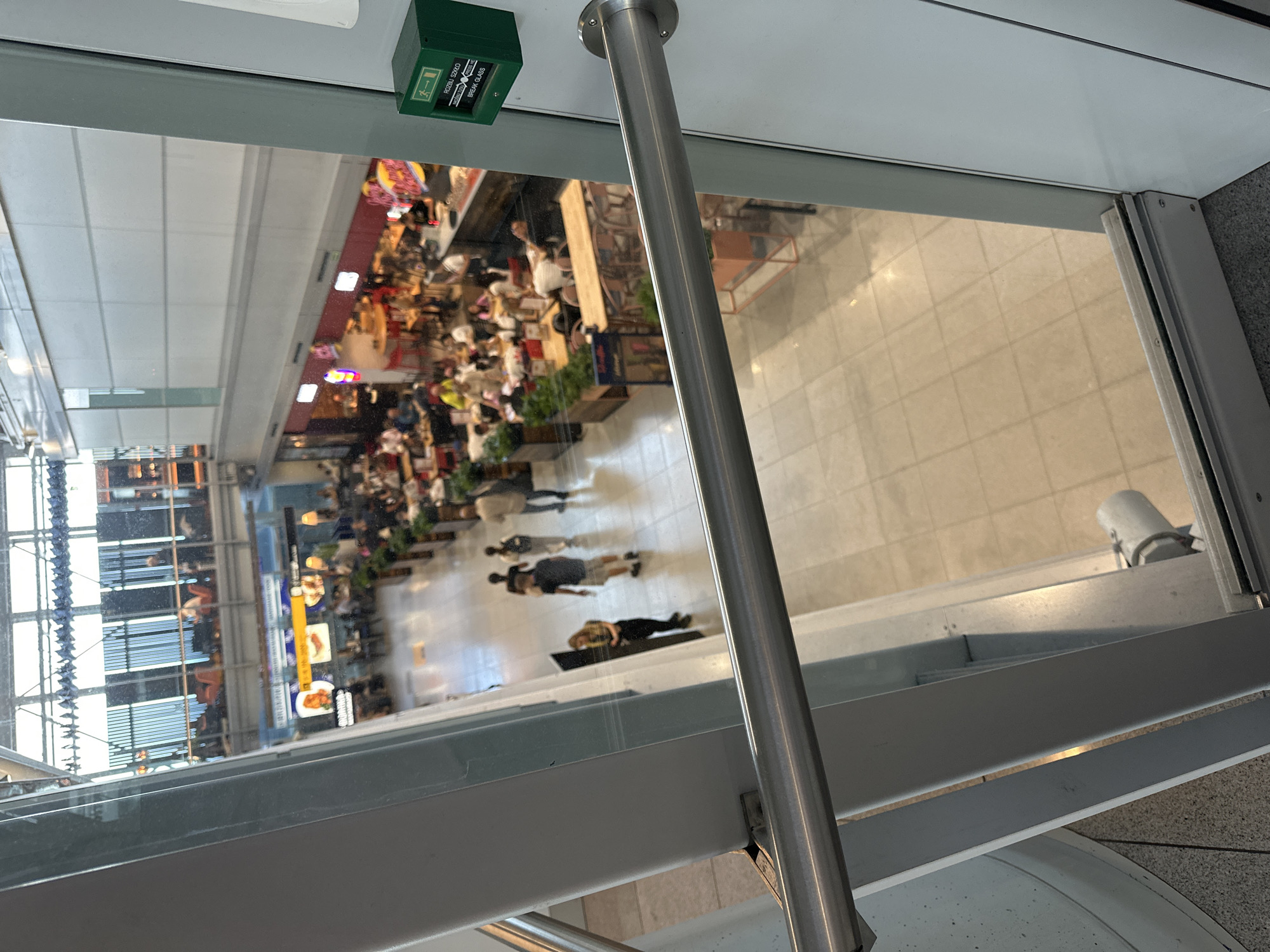 The image depicts an interior view of a busy airport terminal. The perspective is from a higher level looking down onto the main concourse. In the foreground, a metal railing runs horizontally across the frame, suggesting the viewpoint is from a walkway or balcony.
The concourse below is filled with people moving about and seated at various dining establishments. Several restaurants and food stalls are visible, with tables and chairs occupied by patrons. One establishment has a red facade with bright neon signage. People are walking in different directions, some in small groups, others alone.
In the midground, a clear sign with Polish text "ROZBIJ SZKLO" and "BREAK GLASS" is mounted on a green emergency call box, indicating this is likely an airport in Poland. The floor of the terminal is tiled with a light-colored pattern. Plants in planters are strategically placed throughout the concourse, adding a touch of greenery.
The upper levels of the terminal are visible in the background, featuring windows that let in natural light. The overall impression is one of a bustling, modern airport environment. Given the presence of Polish text on the emergency call box, the location is likely Warsaw, Poland.