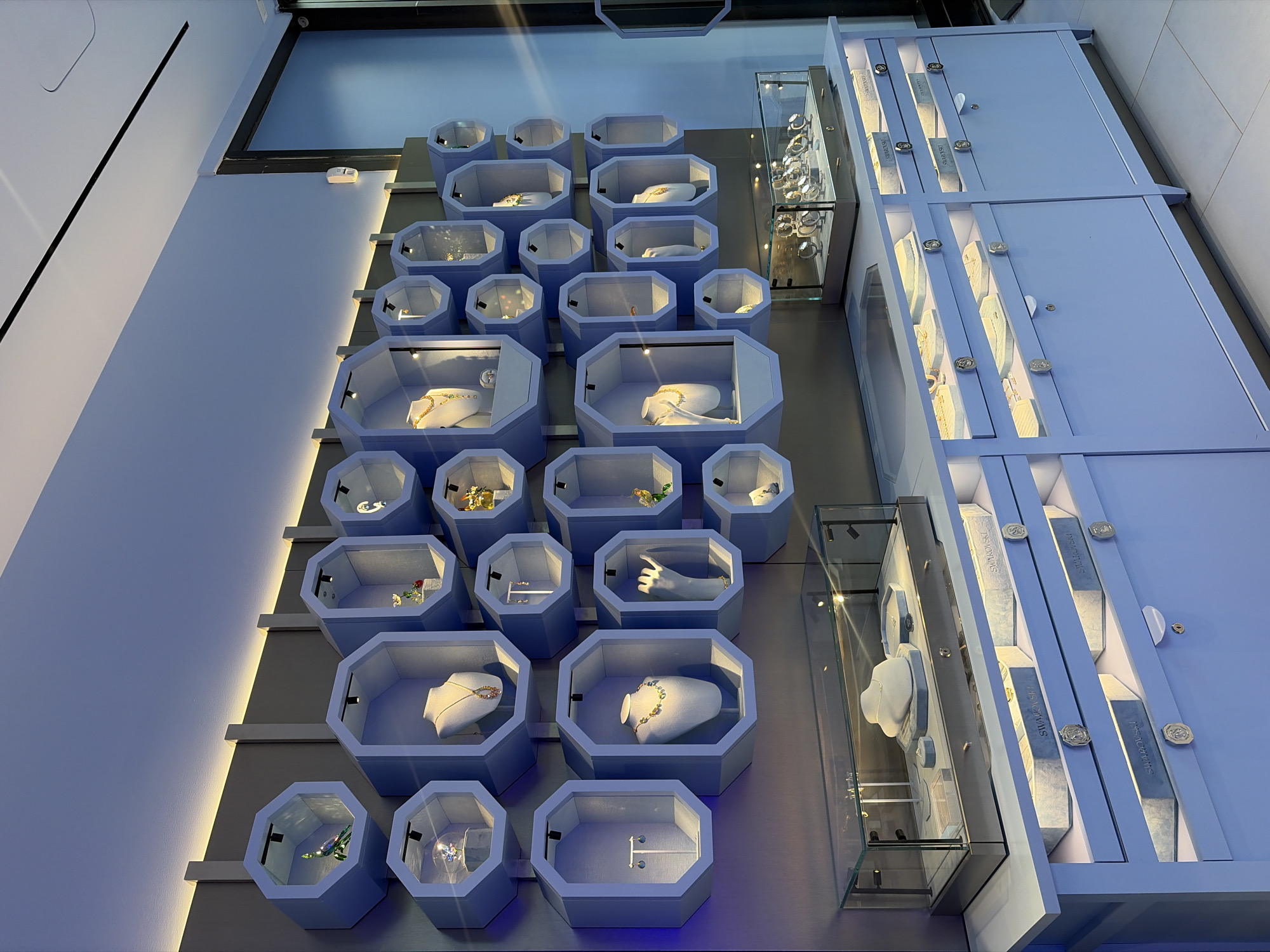This image depicts a jewelry store display in Montreal, Canada. The store features a wall unit with numerous hexagonal display shelves, each lit from above and containing various jewelry items or decorative pieces. These shelves are arranged in a staggered pattern. Some shelves hold necklaces displayed on mannequins, others showcase earrings, rings, and decorative figurines, such as a bird. Below the wall unit, there are glass display cases and drawers. One glass case on a counter displays an assortment of watches. The drawers beneath are a light blue color with decorative silver pulls and some are labeled with "SWAROVSKI". The lighting in the store is soft and ambient, with a strip of warm light running along the top of the wall unit. The overall impression is one of a high-end, modern jewelry boutique.
