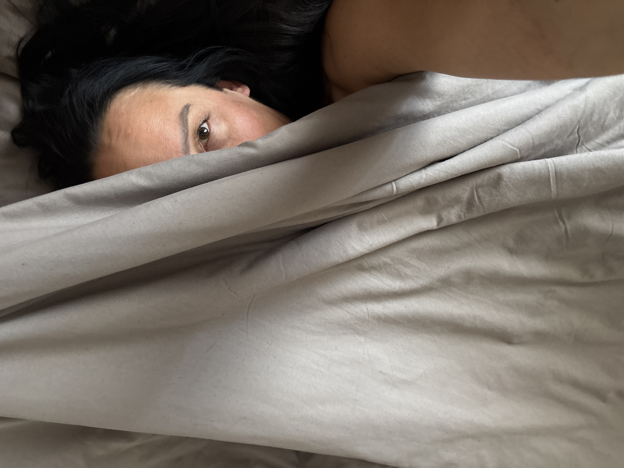 A woman with dark hair is partially concealed under a gray blanket. Only one eye is visible. The scene appears to be indoors, likely a bedroom, with the focus solely on the woman and the blanket. There is no visible information to indicate that the location is Chaikovsky, Russia.