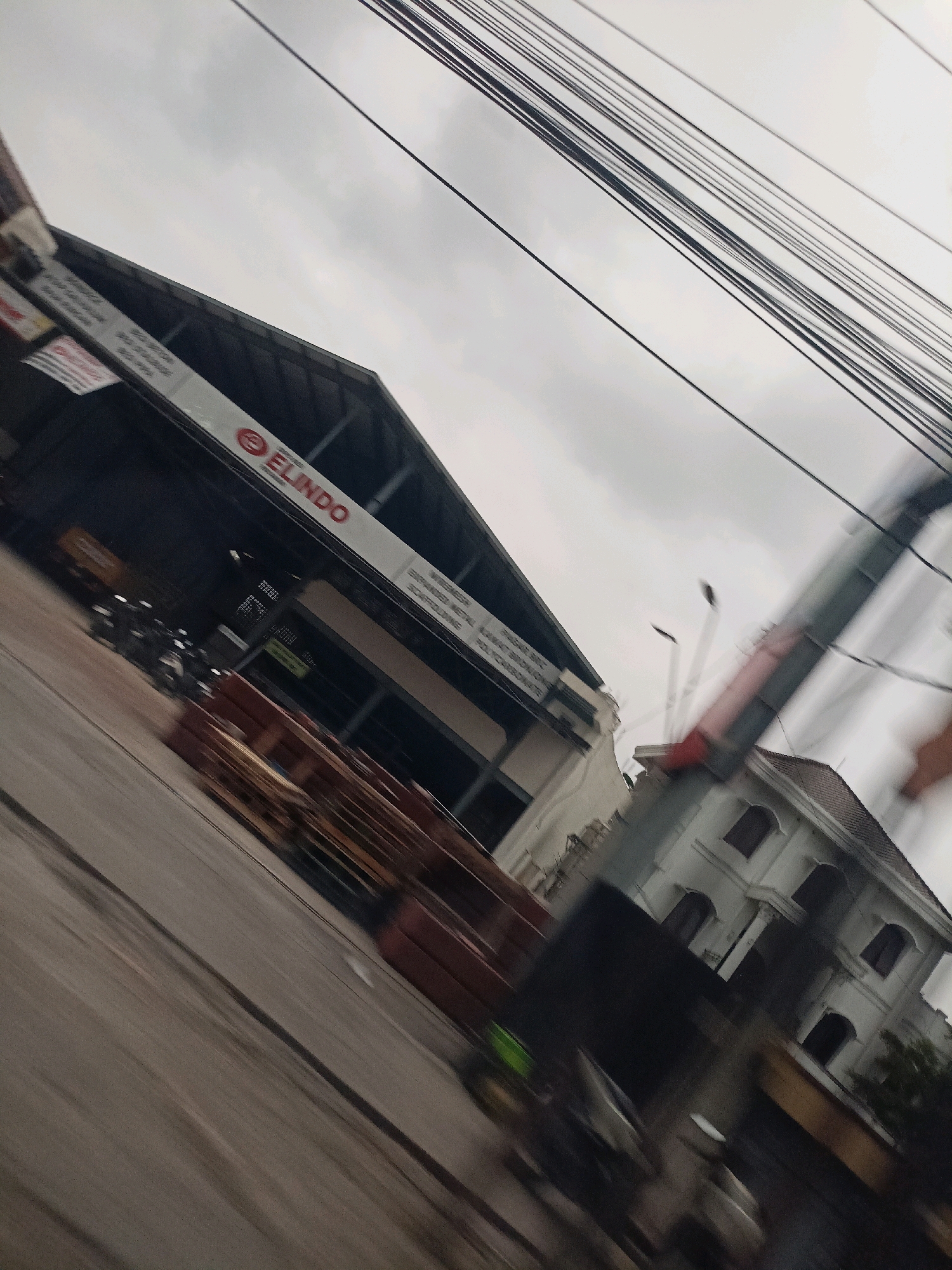 The image, captured in the city of Payaman, Indonesia, depicts a street scene under an overcast sky. A large, dark-roofed building with a prominent "ELINDO" logo in red is visible on the left side of the frame. The building appears to be a commercial establishment, possibly a warehouse or factory, with stacks of brown materials, likely construction supplies, piled in front. To the right of this building, a white, multi-story structure with arched windows is seen. Power lines crisscross the sky, creating a dense network against the gray clouds. The image is partially blurred, suggesting motion, perhaps from a moving vehicle. There are no discernible individuals or specific actions taking place. The overall impression is a glimpse of an industrial or commercial area in an Indonesian city.