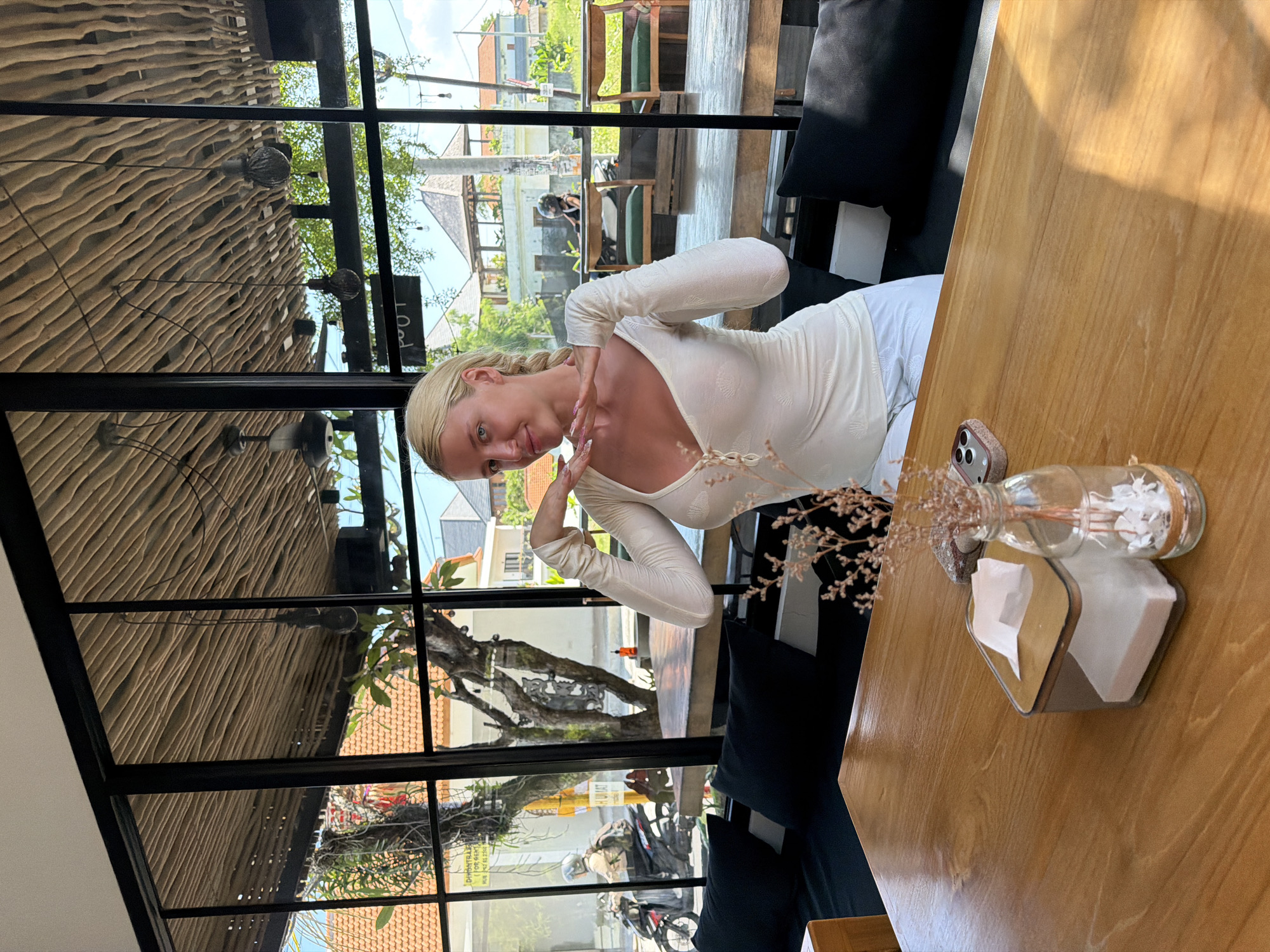 A blonde woman with her hair in a braid sits at a wooden table in a cafe. She is wearing a cream-colored long-sleeved shirt and white shorts. The cafe has large windows that look out onto a street with buildings and trees. There is a scooter with a rider visible outside the window. On the table in front of the woman are a tissue dispenser and a small glass vase with dried flowers. The ceiling of the cafe is made of natural wood beams. The location is implied to be North Kuta, Indonesia.