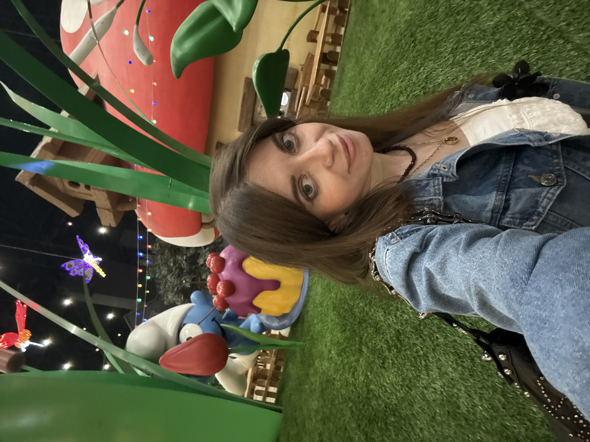 The image presents a selfie of a woman with long brown hair wearing a denim jacket and a white top, captured in what appears to be an indoor entertainment area. She is looking at the camera.
The background features whimsical, oversized cartoon-like decorations, including a red mushroom, a blue and white character with a red nose, a purple and yellow cupcake, and large green blades of grass. There is also what appears to be a birdhouse and light fixtures in the shape of butterflies and dragonflies. Artificial green turf covers the floor.
Based on the subject matter, the location is likely to be a children's play area or a themed entertainment venue in Moscow, Russia, though this is not conclusively determinable from the image alone.