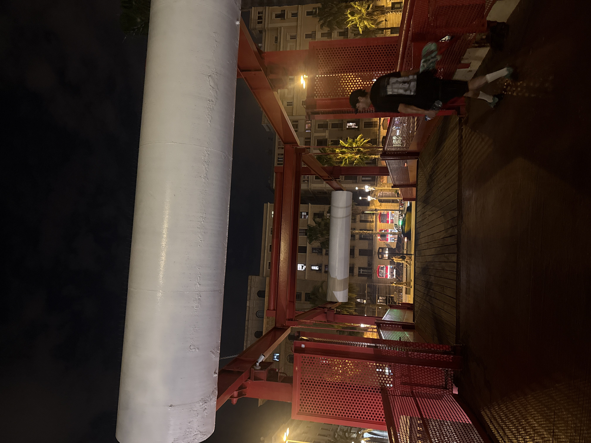 The image depicts an elevated walkway in Barcelona, Spain, at night. The walkway has a red metal frame with perforated panels and wooden decking. A cylindrical white structure stands adjacent to the walkway. A person wearing a black shirt, shorts, and white socks is sitting on the edge of the walkway. The background reveals buildings with lit windows and palm trees. Streetlights illuminate the scene.