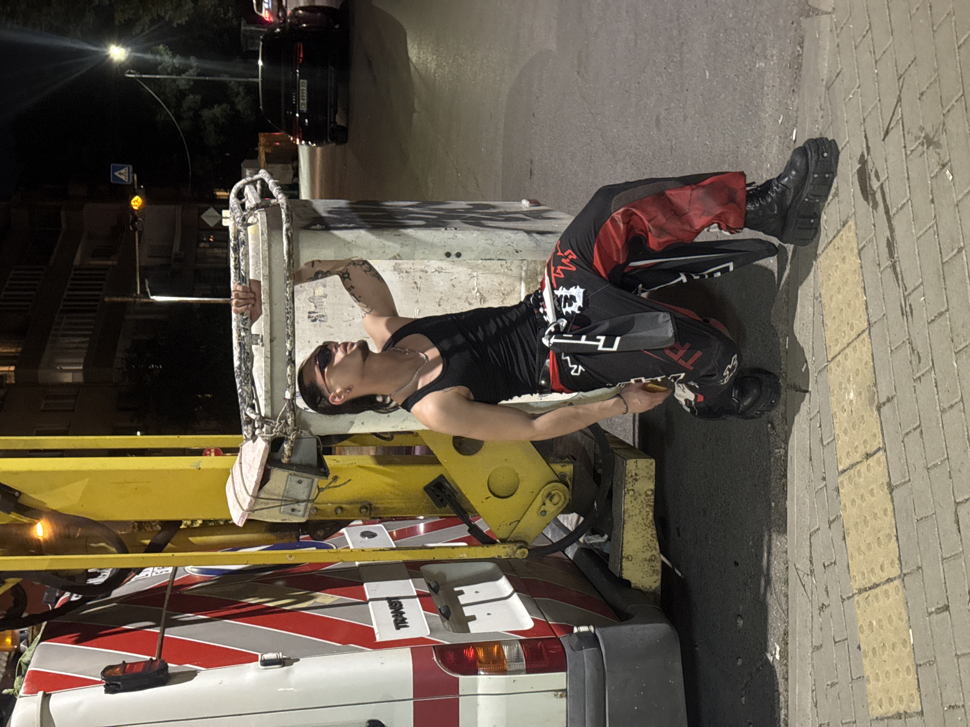 In Sofia, Bulgaria, on a street at night, a young person with tattoos and sunglasses, wearing a black tank top and red and black pants, leans against a dumpster. The person is holding the rim of the dumpster with their left hand. To the left of the dumpster is a white and yellow sanitation vehicle. In the background, a black Audi car is parked on the street, and buildings are visible with streetlights illuminating the area. The ground is paved with cobblestones and tactile paving blocks.