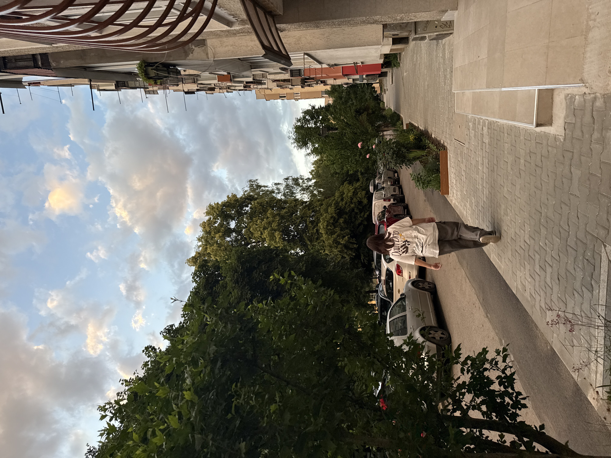 A person wearing a white t-shirt with a graphic design and gray pants walks away from the camera down a sidewalk. The person is on the right side of the image. To the left of the sidewalk is a street with parked cars, including a silver Volkswagen Golf and a white car. Trees line the street and sidewalk, with lush green foliage. On the right side of the image, a multi-story building with balconies and windows faces the street. The sky is cloudy with patches of blue and the sun is low, suggesting it is either early morning or late afternoon. The scene appears to be in a residential area of Sofia, Bulgaria, likely a courtyard or street between apartment buildings.