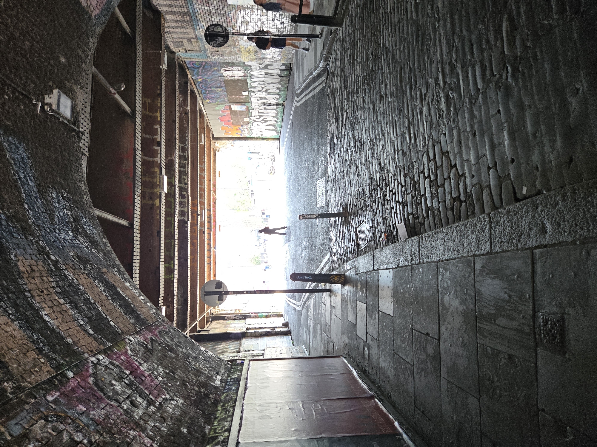 The image depicts an urban alleyway in London, United Kingdom, characterized by a combination of textures and structures. The left side features a curved brick wall, possibly a viaduct, adorned with graffiti and showing signs of weathering. Parallel to it is a structure with vertical beams and horizontal supports, creating a sense of enclosure. The right side of the alley consists of cobblestone pavement leading to a smoother, tiled walkway. Two bollards are visible in the foreground and midground, each with a circular sign mounted on top. The background shows figures walking toward the brightly lit end of the alley, suggesting an exit point. The overall environment is dimly lit, with natural light entering from the opening at the end. The alley exhibits a mix of decay, urban art, and functional infrastructure.