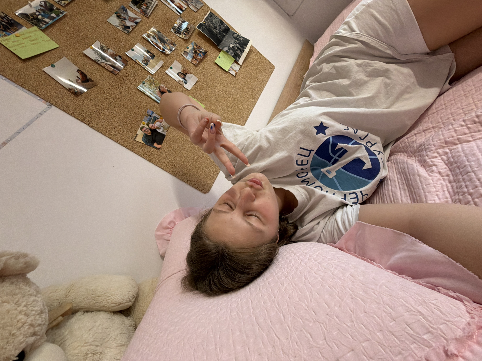 A young person is lying on a bed with pink bedding, captured from an elevated, slightly tilted perspective. The individual, with brown hair, appears relaxed with their eyes closed and lips slightly puckered. They are wearing a light-colored T-shirt with a blue and white graphic design and white shorts. Their right arm is raised, forming a peace sign with their fingers, which have blue and white nail polish. A light-colored bracelet is visible on their wrist.
The T-shirt prominently displays text in Cyrillic script, including "МОРЕЦ" (MORYETS) and "БУРГАС" (BURGAS), along with a star and a nautical-themed graphic resembling an anchor or boat. This text directly references the city of Burgas, Bulgaria.
Behind the person's head, a cork board is affixed to a white wall, covered with numerous small, pinned photographs, suggesting personal memories or moments. A small green sticky note with illegible handwriting is also pinned to the board. To the left of the bed, several light-colored stuffed animals, resembling teddy bears, are visible. The overall lighting suggests it is daytime. The scene conveys a casual, personal, and relaxed atmosphere, likely in a bedroom.