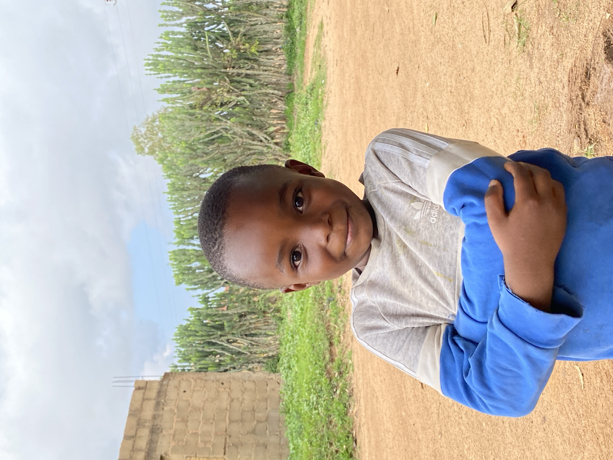 A young boy stands outdoors in Kuru Dungus, Nigeria. He has short, dark hair and wears a light grey, long-sleeved shirt with blue accents. His right hand rests on his left arm. The background features a dirt ground, green vegetation, trees, and a partially constructed cinder block building. The sky is visible and overcast.