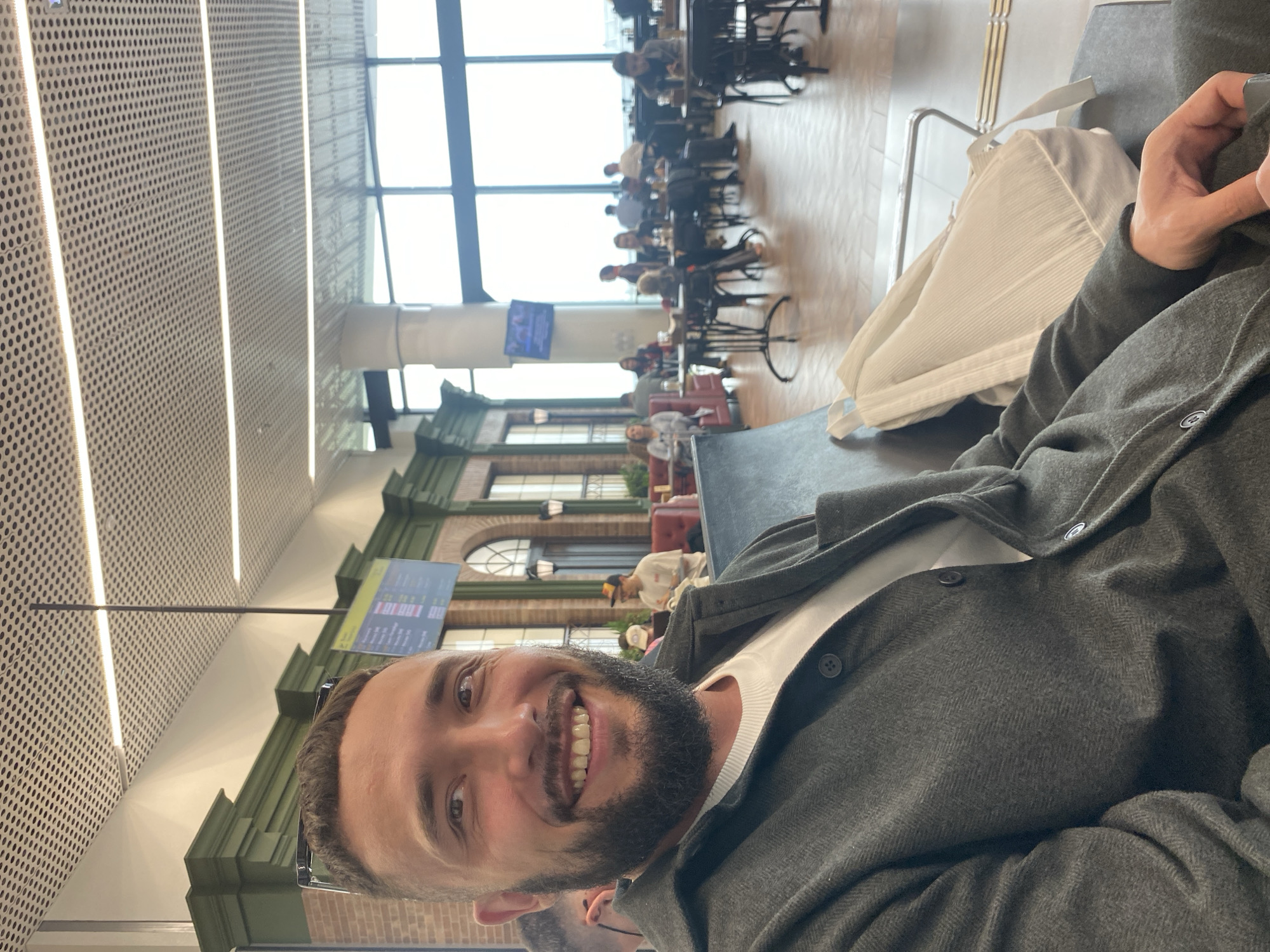 The image captures a man with a beard, wearing a grey jacket over a white t-shirt, smiling at the camera. He is seated in what appears to be an airport waiting area, possibly in Новое Мартьяново, Russia, indicated by the presence of seating, a departure board in the background, and large windows offering a view of the outdoors. Other individuals are visible in the background, seated at tables in a dining or lounge area. A white bag is placed next to the man on his seat. The ceiling features a modern design with perforated panels and integrated lighting. The overall environment suggests a public space within a transportation hub.