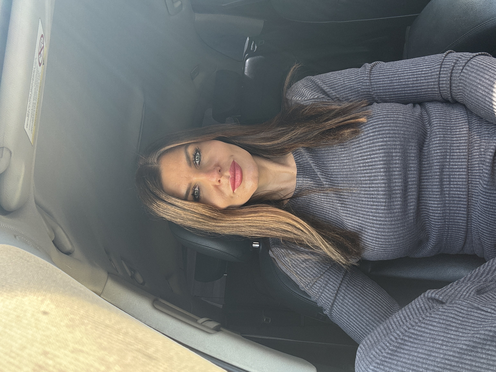 A woman with long brown hair and grey eyes is sitting in the passenger seat of a car. She is wearing a grey ribbed sweater. The interior of the car is mostly dark, with the door and visor in the foreground appearing in a light beige tone. The location is unknown.
