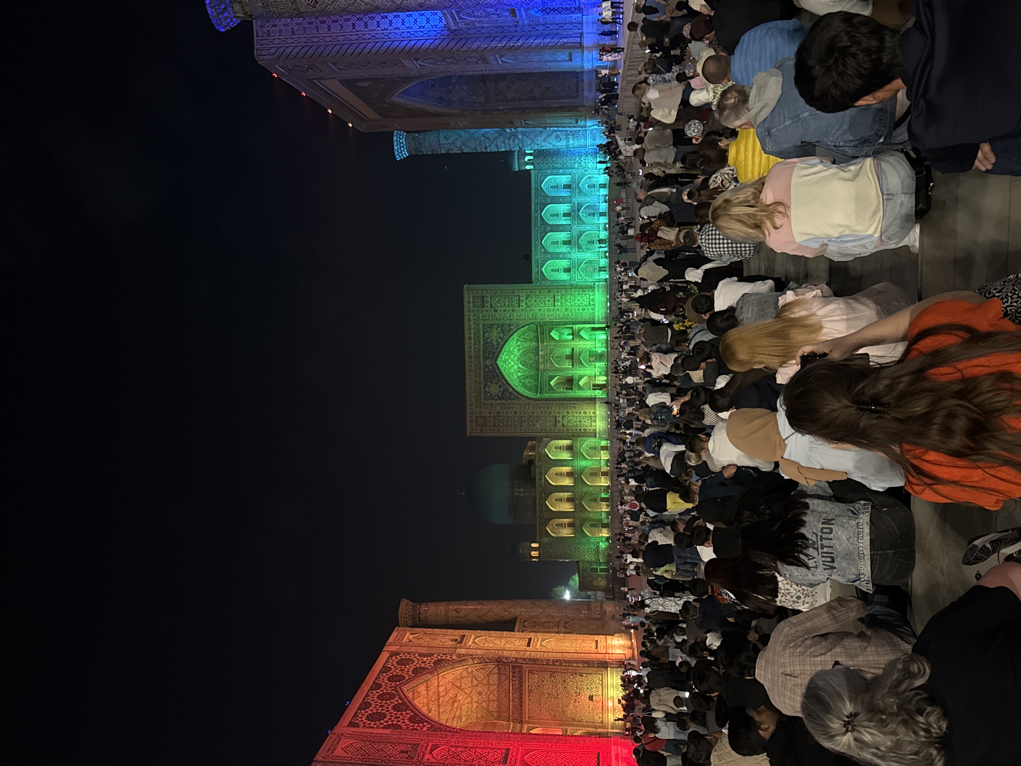 A wide shot at night in Samarkand City, Uzbekistan, shows a large group of people bowing in prayer facing ornate, colorfully lit historical buildings. The people are diverse in age, clothing, and hair color. The buildings are lit up in vertical color blocks, beginning with red at the bottom, shifting to yellow, green, blue, and purple at the top. The background is black. The composition is vertically oriented.