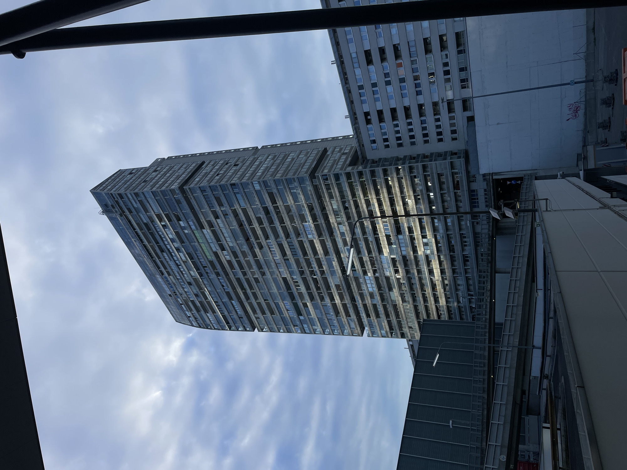 View of a Brutalist-style building under a cloudy sky in an unknown city. The building is composed of multiple horizontal sections of concrete and glass, with many small windows. Another similar building is visible on the right side of the frame. The perspective is from below, looking up at an angle. Portions of other urban structures are visible at the bottom and to the side.