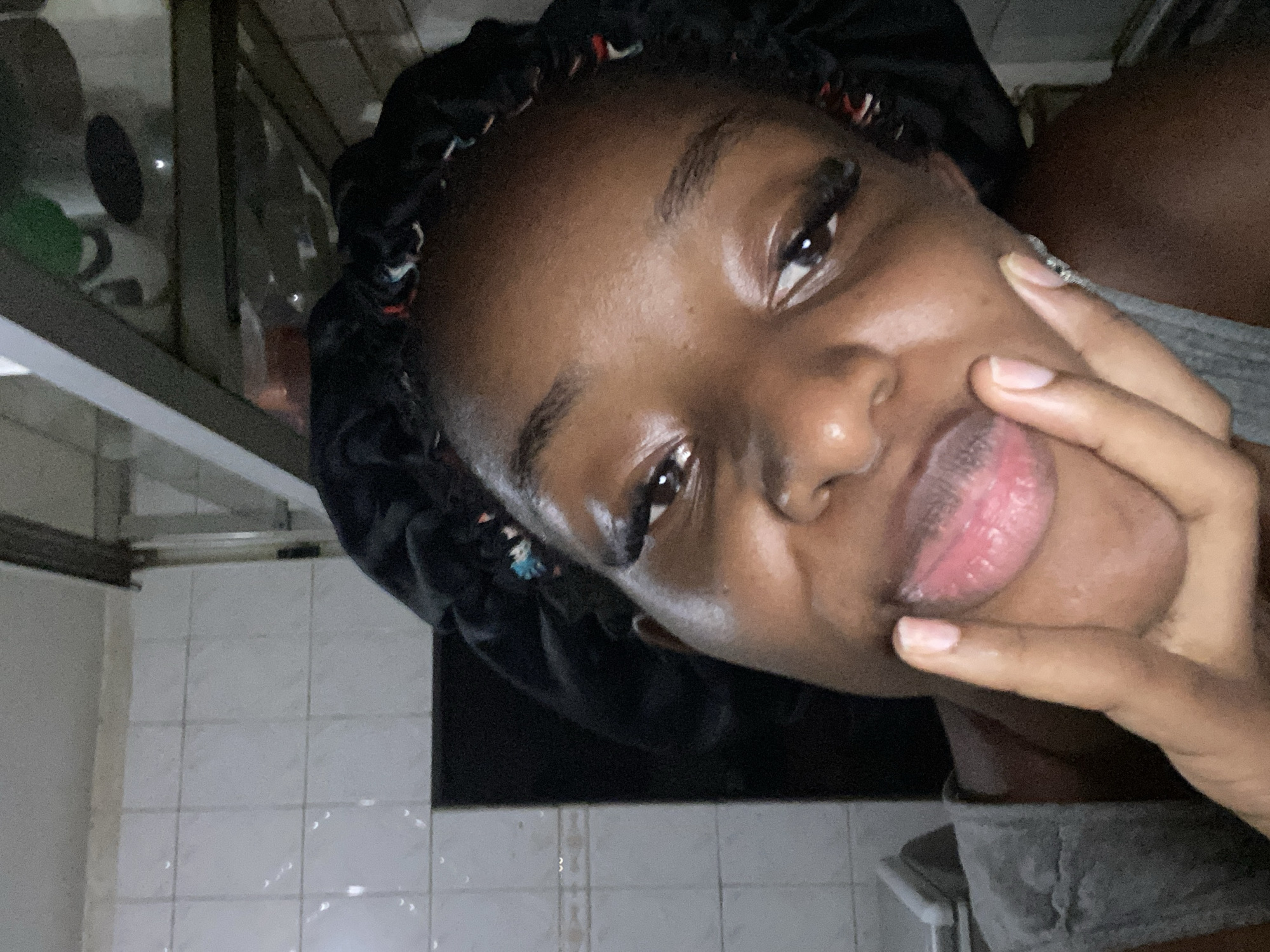 The image shows a close-up, frontal view of a young Black woman wearing a black bonnet. Her hand is placed on her chin, and she is looking towards the camera. The background appears to be a tiled bathroom or kitchen with white tiles on the wall and a stainless-steel fixture or shelf above. The lighting is dim, creating shadows. The woman has visible makeup, including mascara and possibly lip gloss, enhancing her eyes and lips. The scene does not contain any specific indicators that would allow for the identification of the city of Jos, Nigeria.