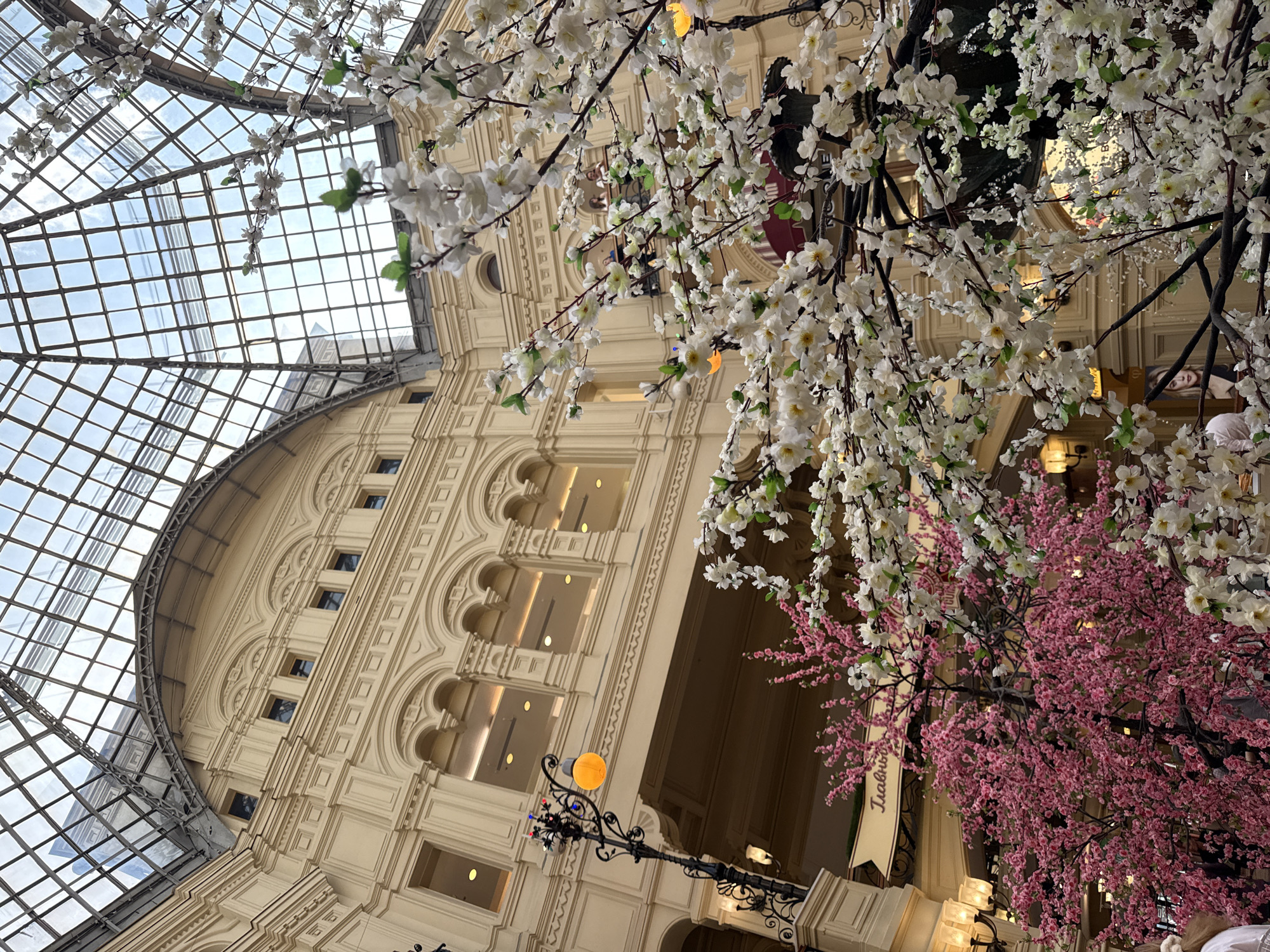 The image shows the interior of GUM (State Department Store), a major shopping mall in Moscow, Russia, located on Red Square. The building's ornate facade is visible, with multiple levels of arched windows and detailed ornamentation. The interior is bathed in soft light filtering through the arched glass ceiling. Decorative flowering trees, both white and pink, are prominently displayed in the foreground, partially obscuring the view of the building. A vintage-style street lamp with an orange globe is positioned near the base of the building. There are people present, but they are indistinct and blended into the background. A banner with Cyrillic text hangs near one of the trees.