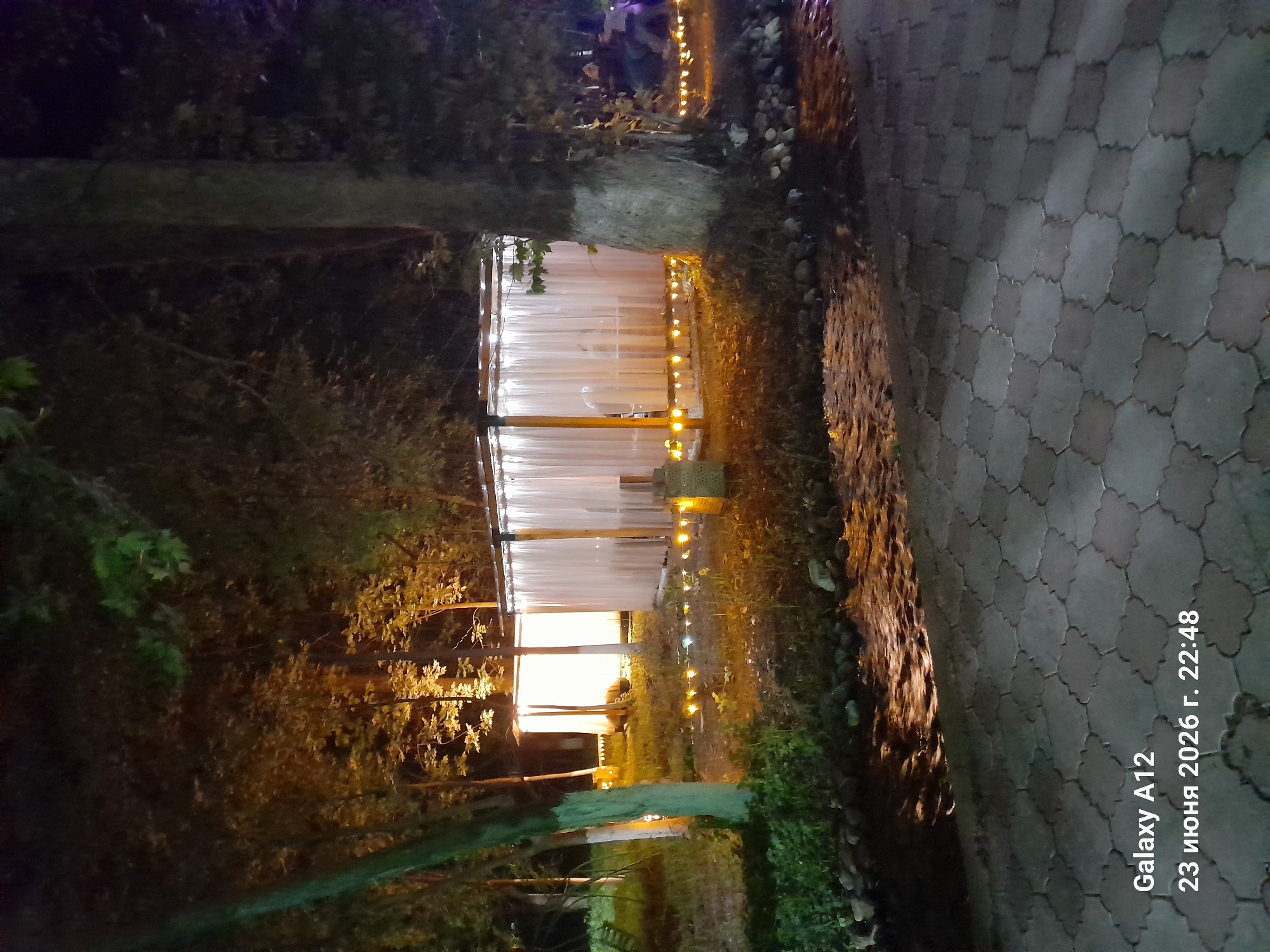 The image depicts a nighttime scene in Kara-Köl City, Kyrgyzstan. The focus is on a structure that appears to be an outdoor seating area or a small pavilion, enclosed by translucent white curtains. Warm yellow lights are strung along the exterior of the structure, creating an inviting ambiance. The area is surrounded by trees and foliage, suggesting a natural or park-like setting. A paved walkway with hexagonal stones is visible in the foreground, partially illuminated by the lights and the dim ambient light of the night. The overall mood is serene and tranquil, enhanced by the soft lighting and the natural elements.
Visible text at the bottom right of the image reads "Galaxy A12 23 июня 2026 г. 22:48", indicating the device used to capture the photo, the date, and the time. The presence of the date "2026" suggests this is a future date. The time is late evening, consistent with the nighttime setting. There are no people visible in the frame, but the setup of the pavilion suggests it might be a place for gatherings or dining. The weather appears calm, with no indicators of rain or wind.